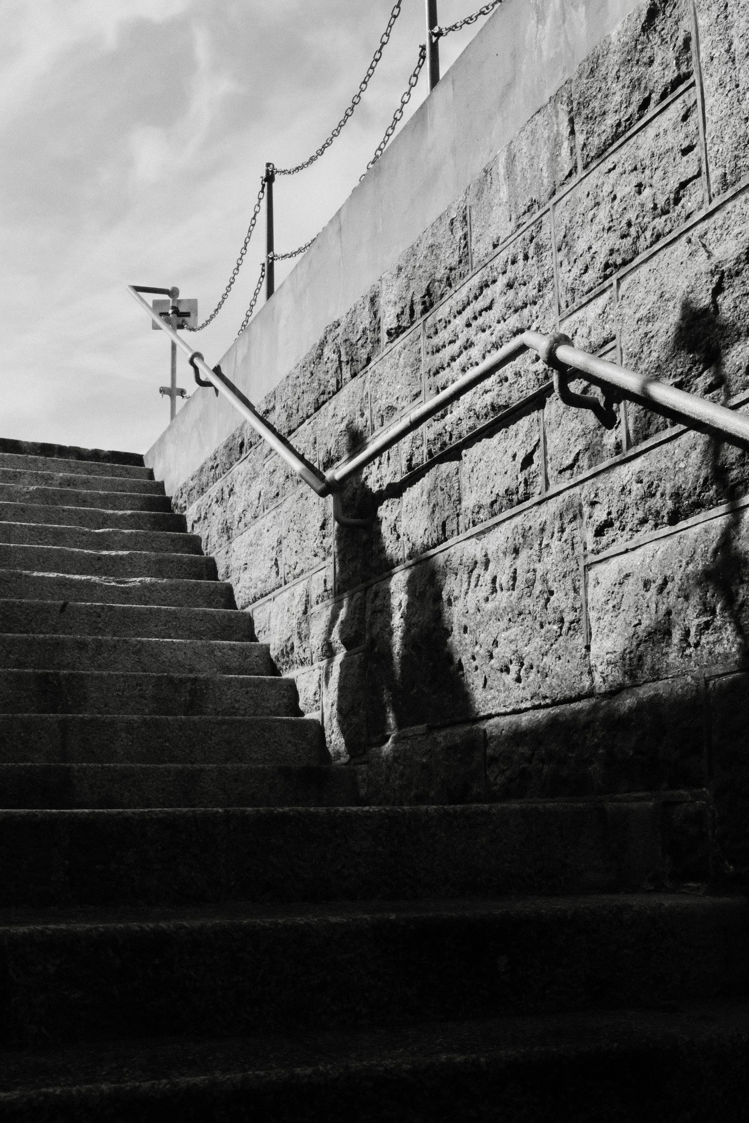 A black and white photo of a staircase with a metal handrail and chain barrier along a stone wall, viewed from the bottom looking up.