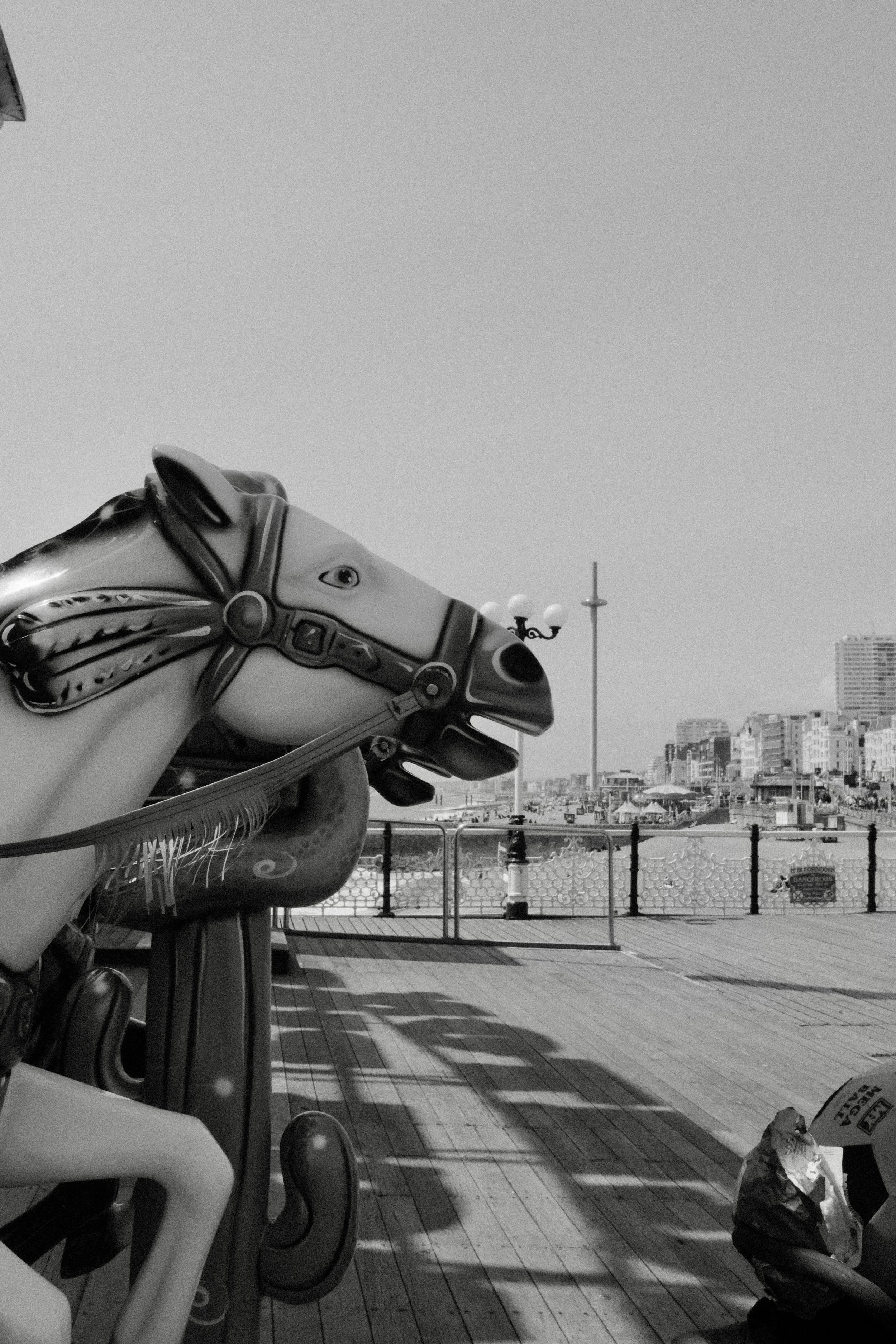 Black and white photo of carousel horse ride on a boardwalk, with a cityscape in the background and a person sitting under an umbrella.