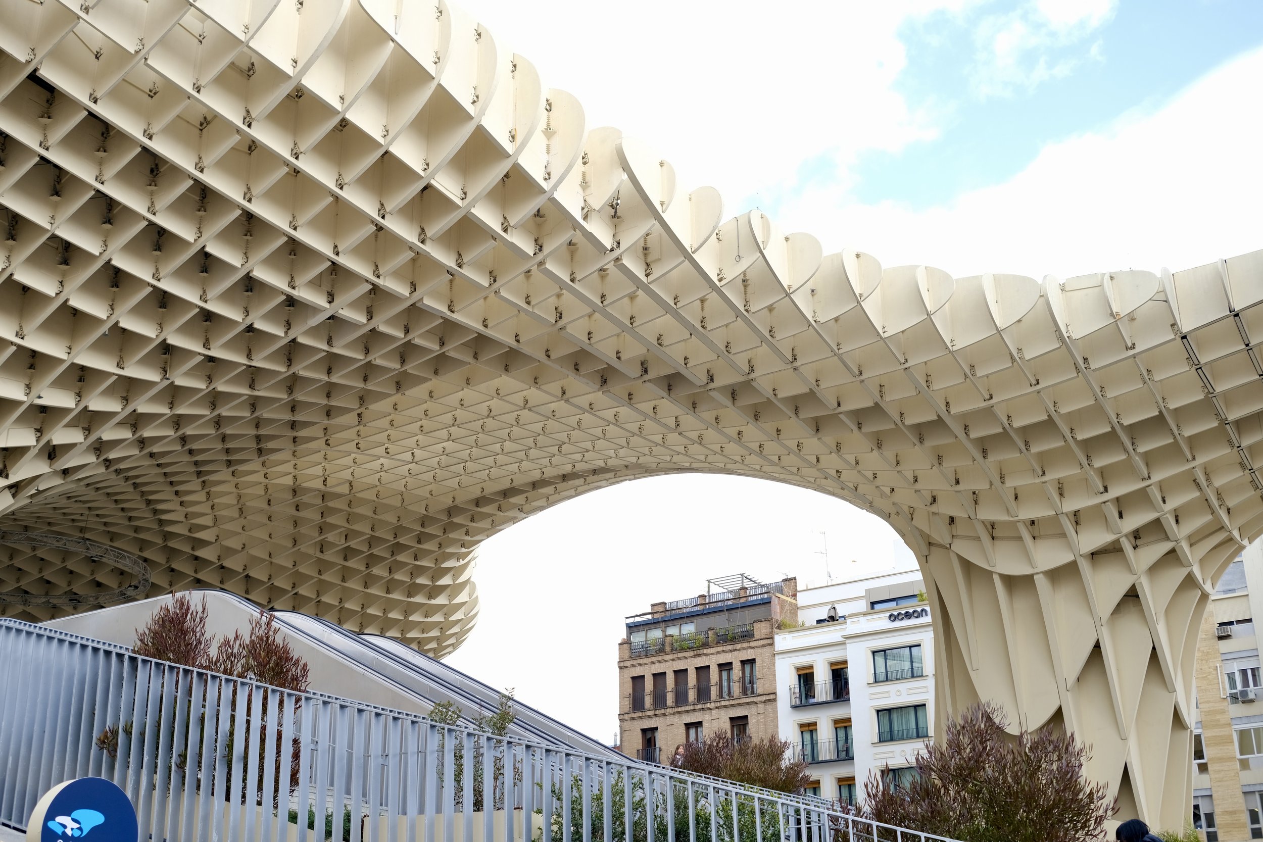 Close-up of architectural structure with a grid-like pattern of white panels supported by metal brackets, set against a blue sky with clouds, in an urban area with buildings and trees nearby.