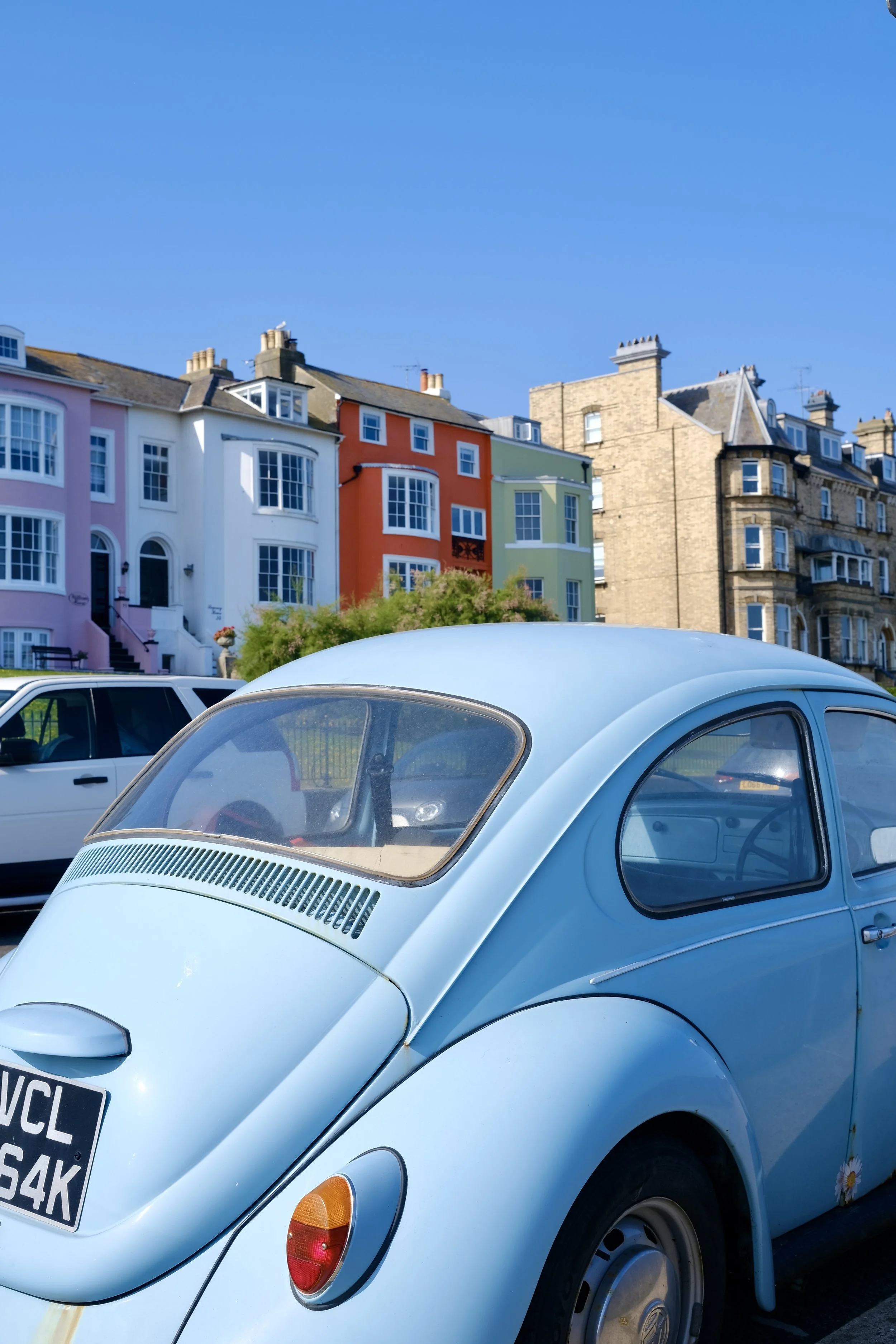 A sky blue vintage Volkswagen Beetle parked on a street, with a row of colorful houses in the background and clear blue sky overhead.