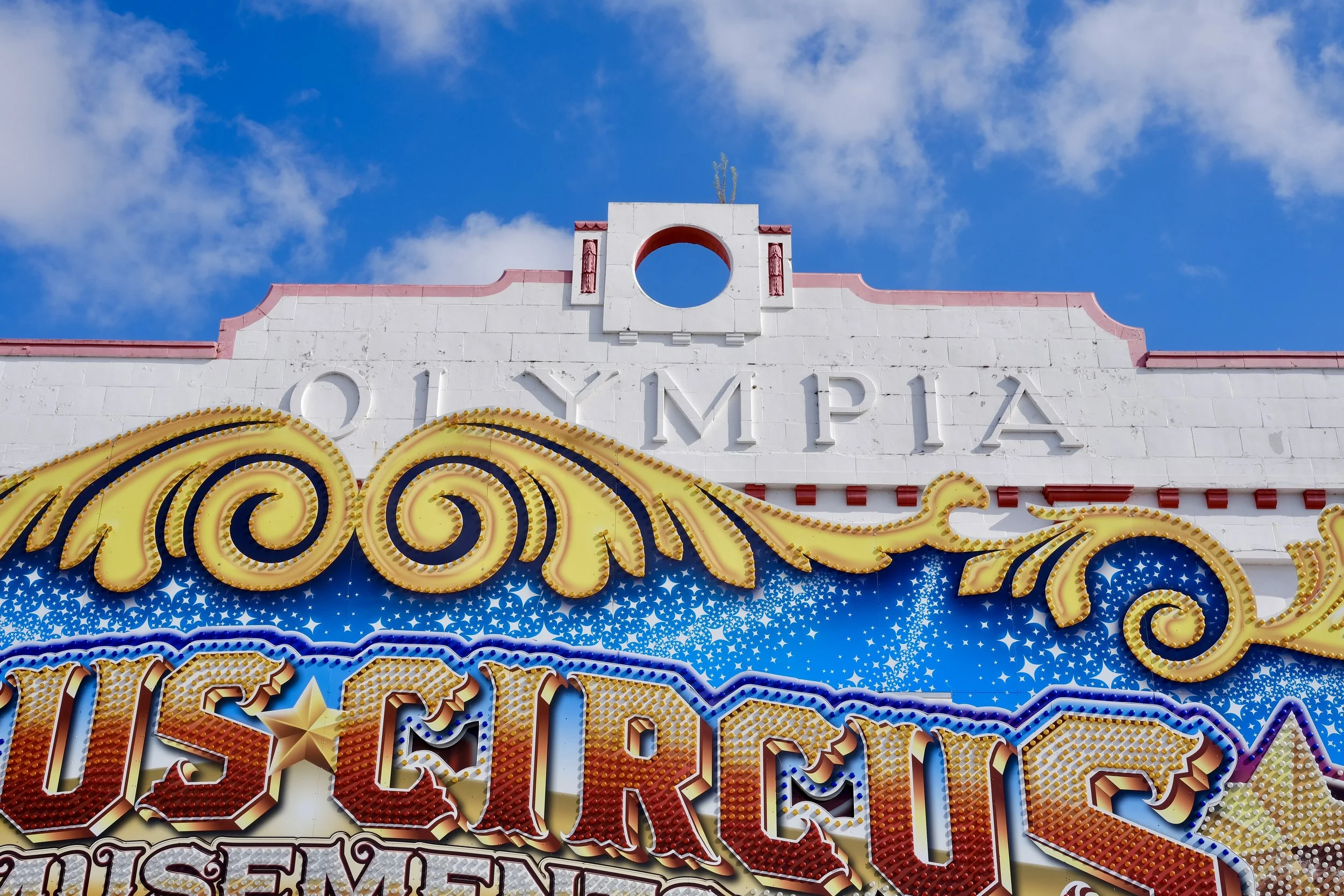 Close-up of a vintage carnival sign with bright lights, featuring the word 'OLYMPIC' on a white building under a partly cloudy sky.