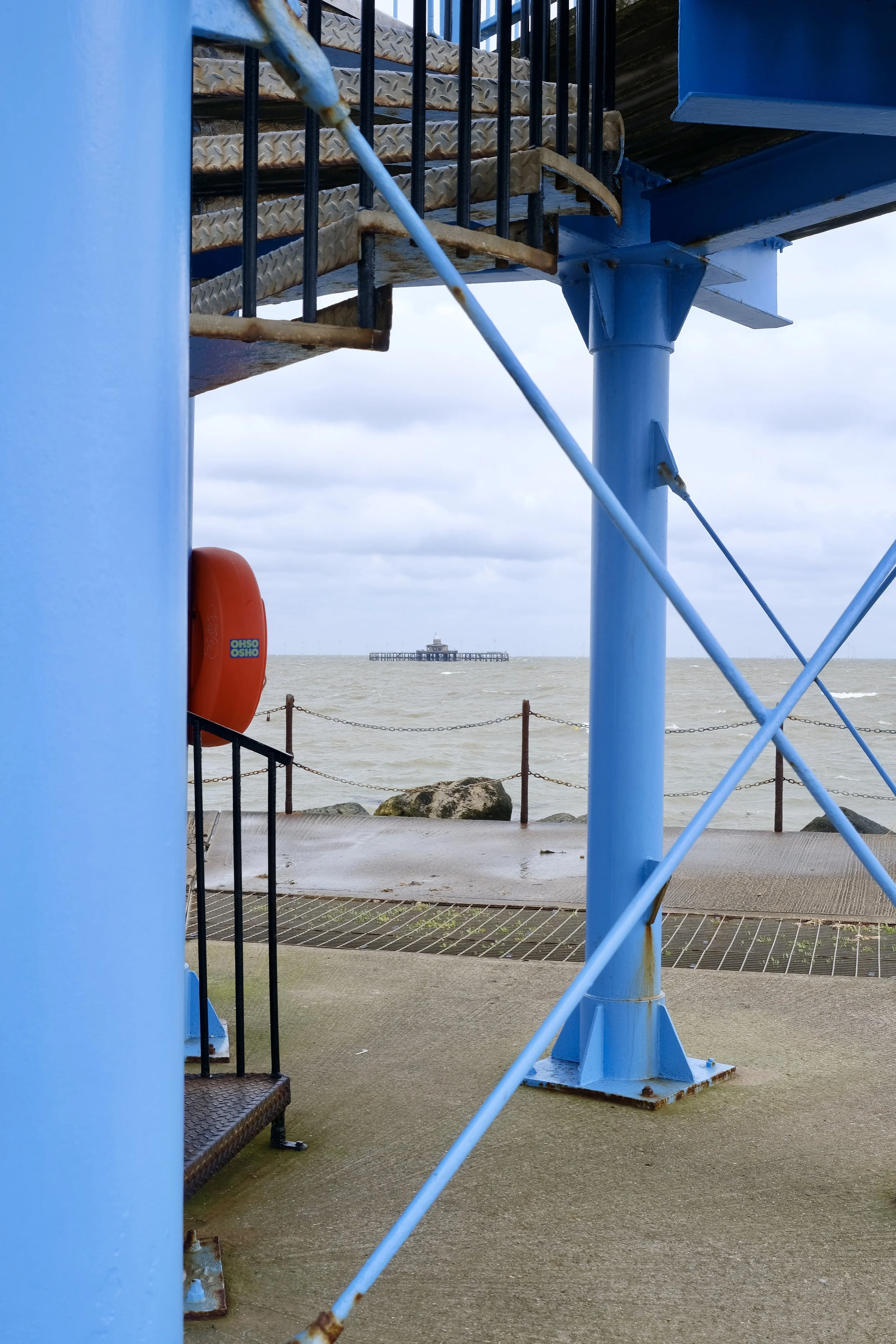 A view from a lifeguard tower looking out towards a pier in the ocean. The tower is painted bright blue, with metal stairs and railings. An orange life preserver is attached to the tower, and a chain fence runs along the edge of the walkway on the be