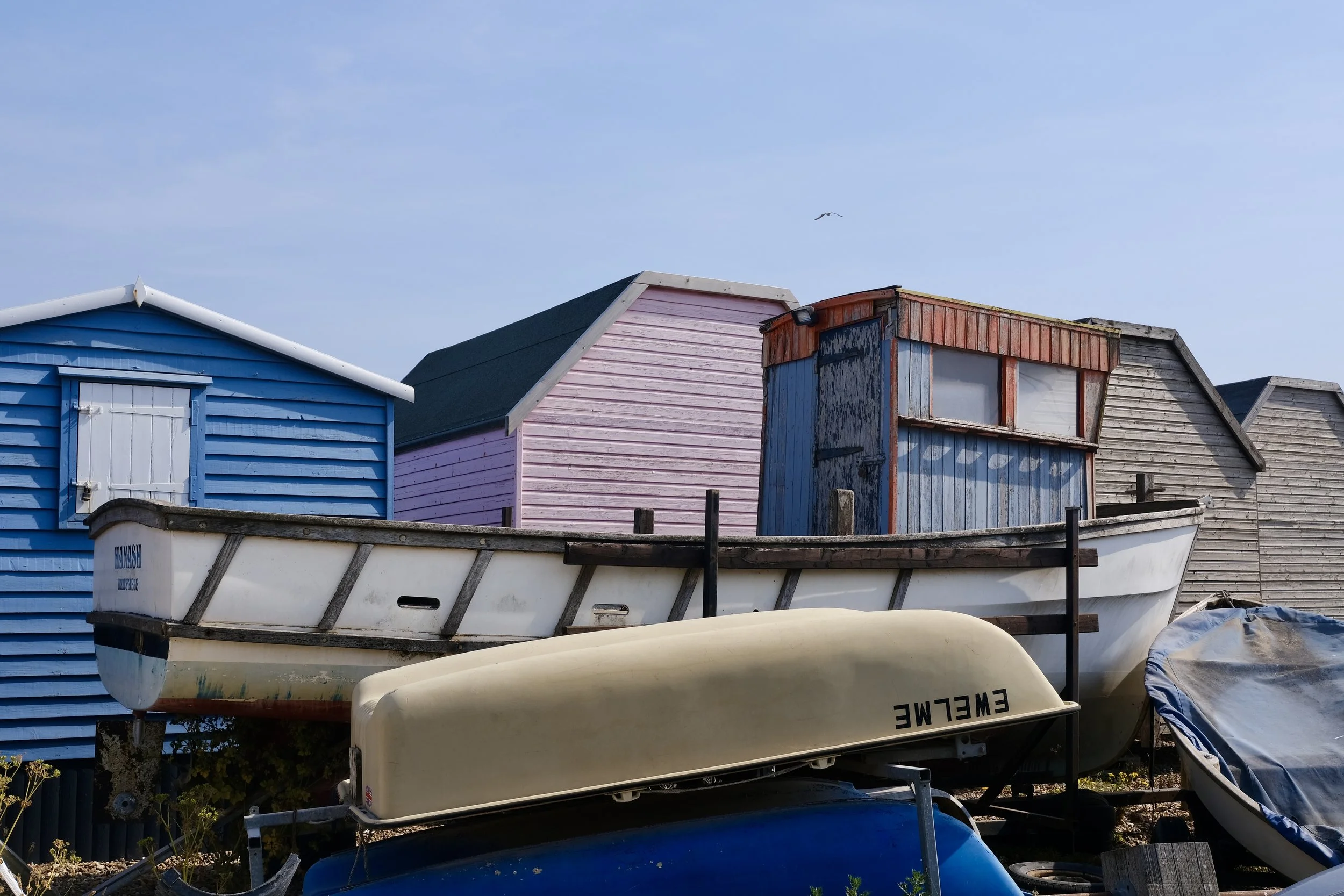 Colorful beach huts in blue, pink, orange, and gray, with boats stored underneath and around them, in a seaside setting.