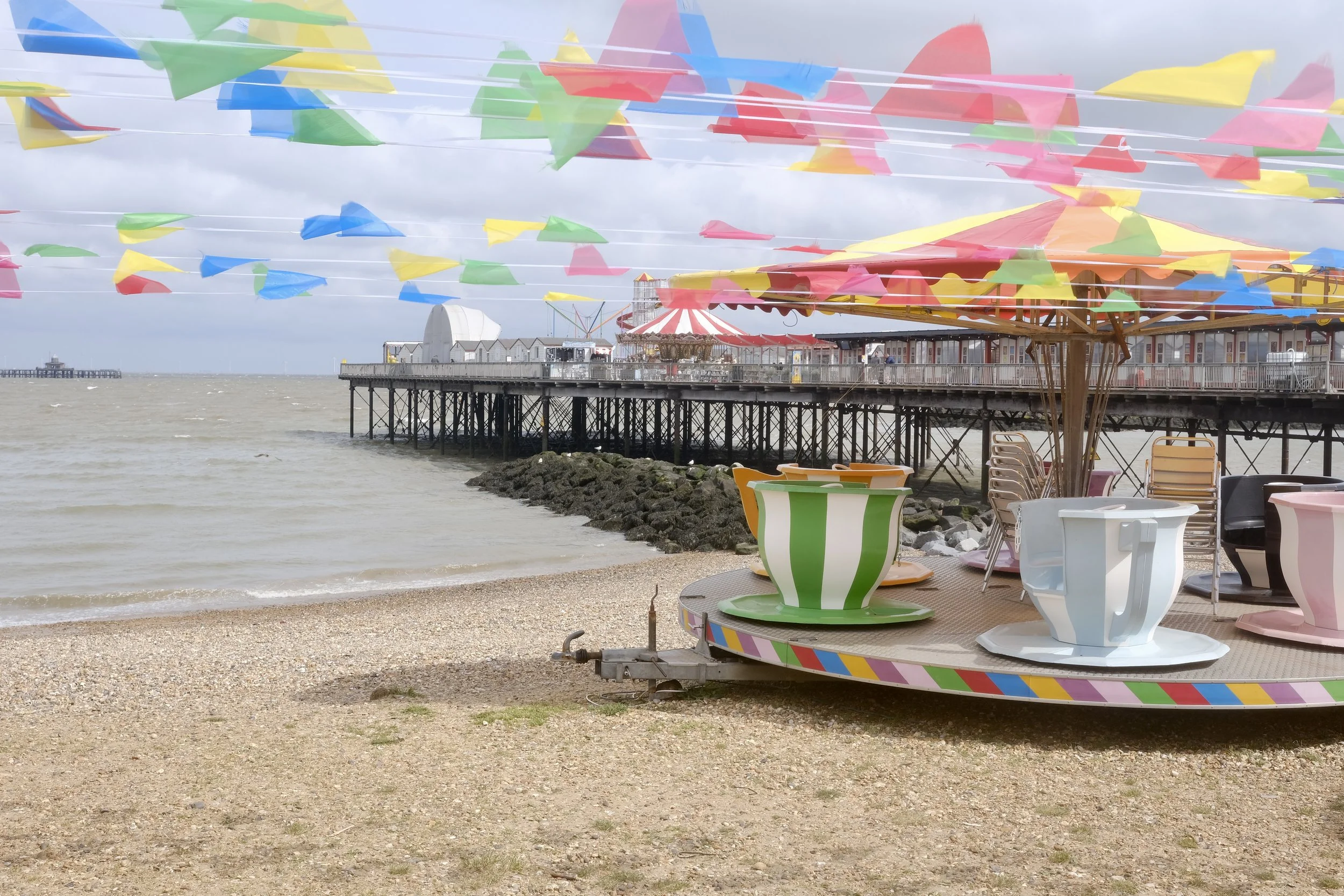 Colorful amusement park teacup ride on a beach with a pier, ocean, and cloudy sky in the background.