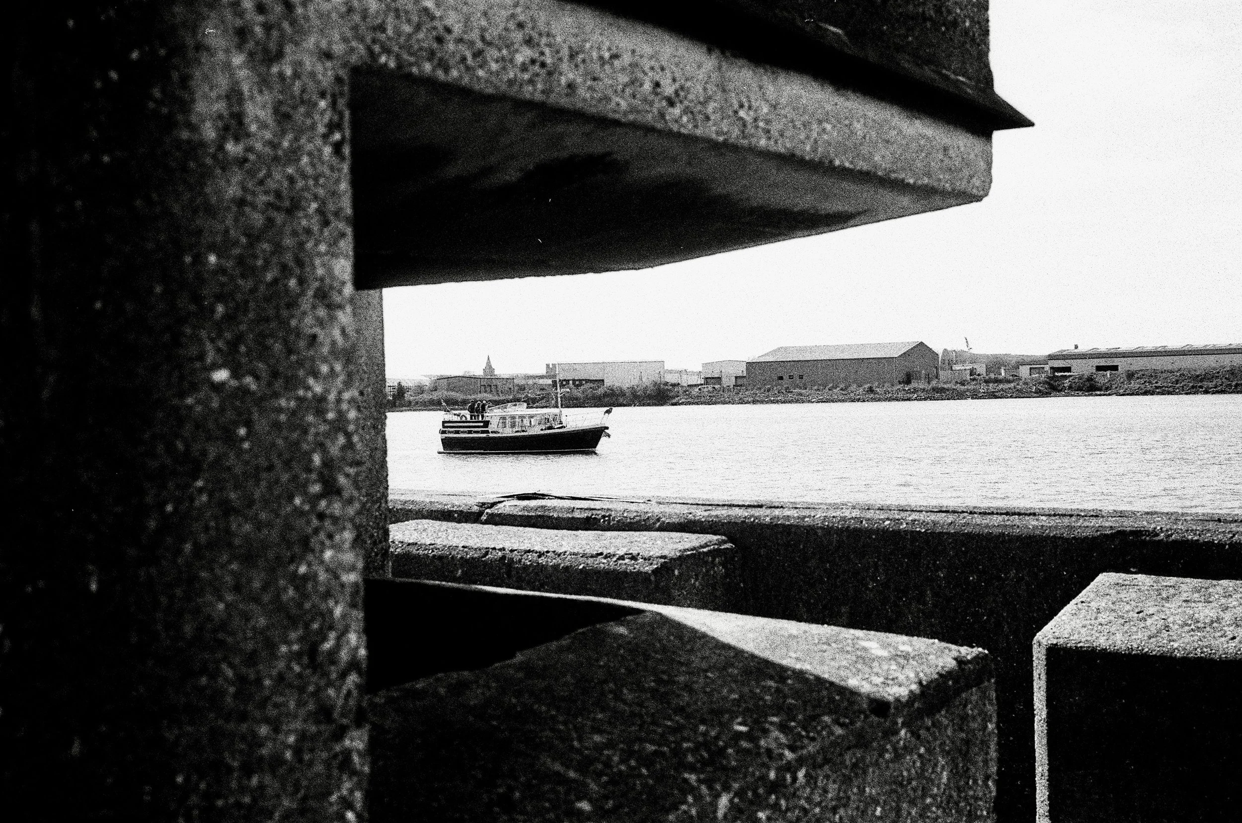 Black and white photo of a boat on a river seen through a gap between two large brick structures.