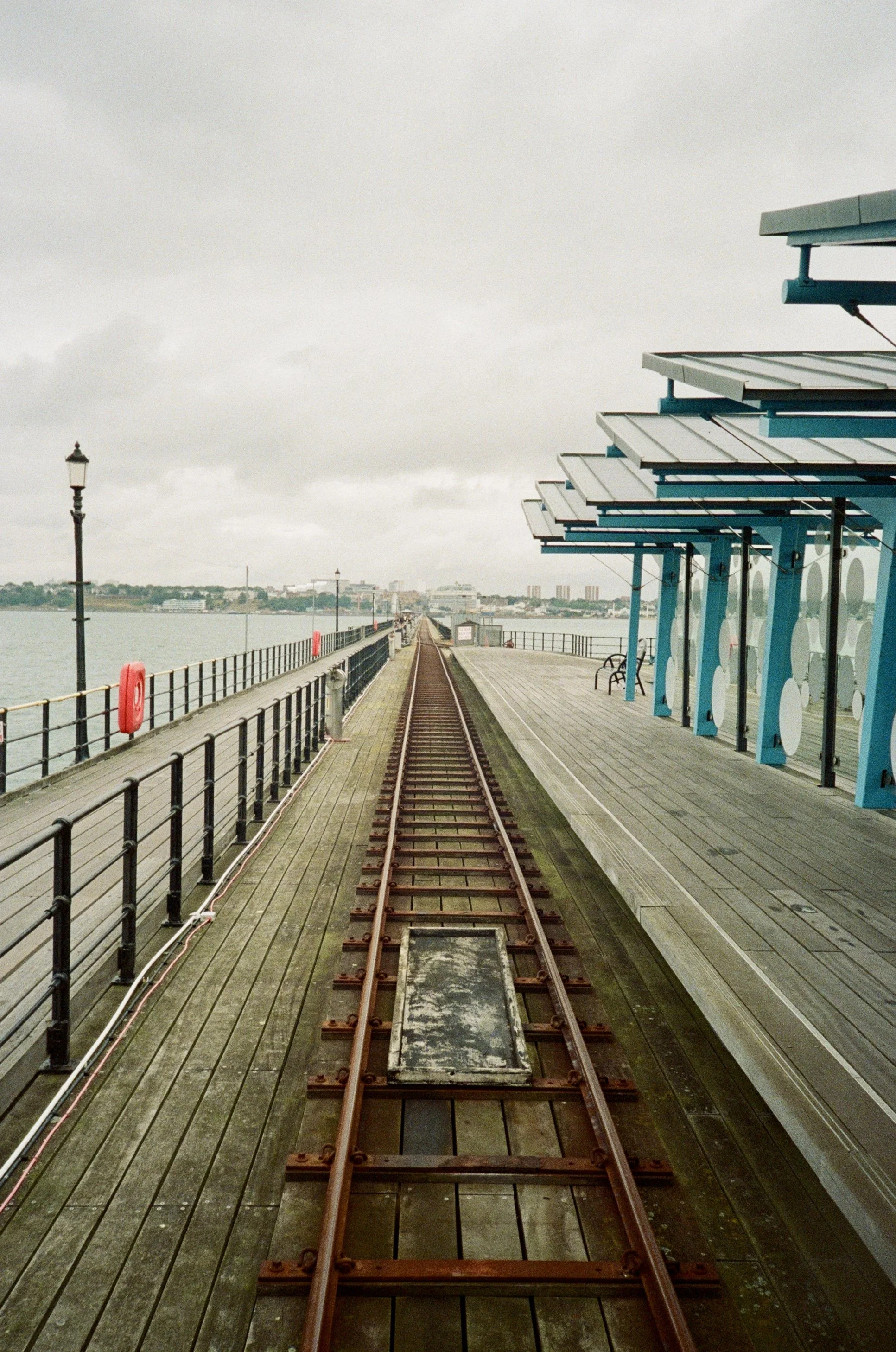 A pier with a rail on the left and a building with a blue canopy on the right. A train track runs down the center of the pier, extending into the distance over water with a cityscape in the background. The sky is cloudy.