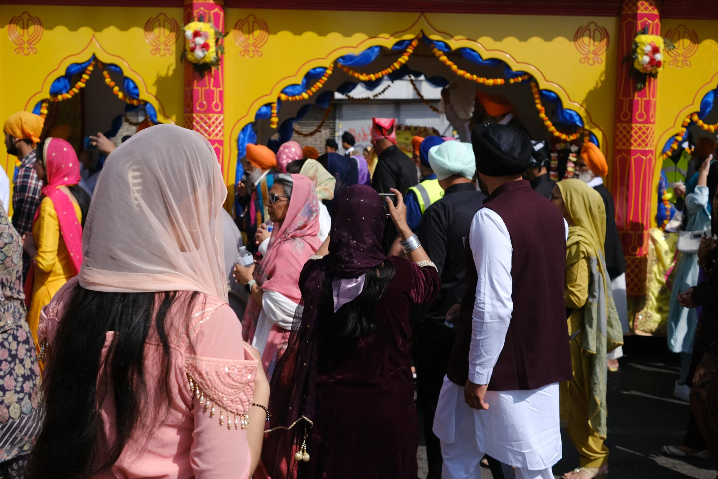 Crowd of people dressed in traditional Punjabi attire, gathered at a festive event with a bright yellow decorated backdrop.