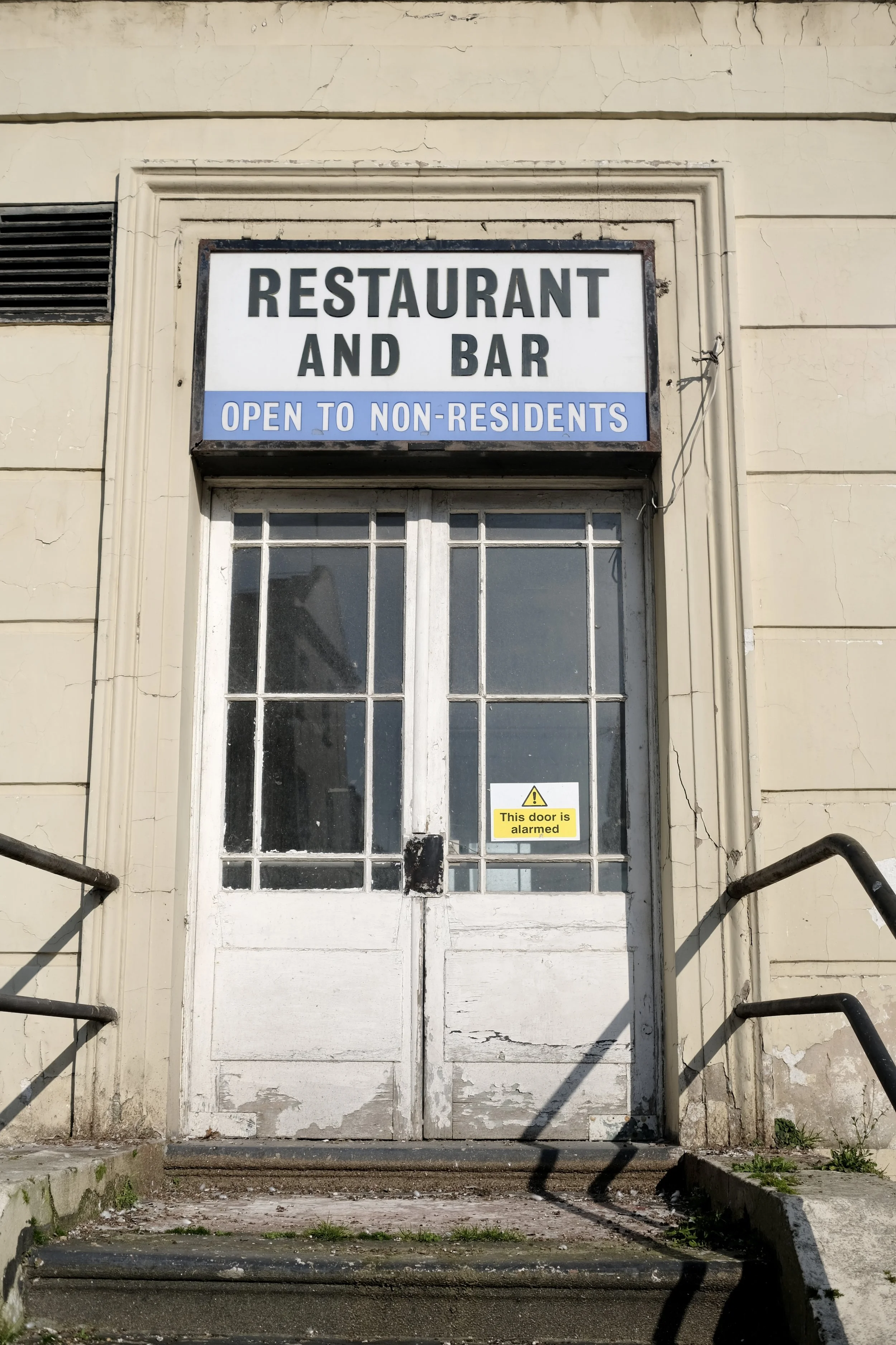 Exterior view of a weathered door with a sign above reading 'RESTAURANT AND BAR OPEN TO NON-RESIDENTS' and a small yellow sticker on the door stating 'This door is alarmed.' The door has a glass window with visible cracks and a small handle, flanked 