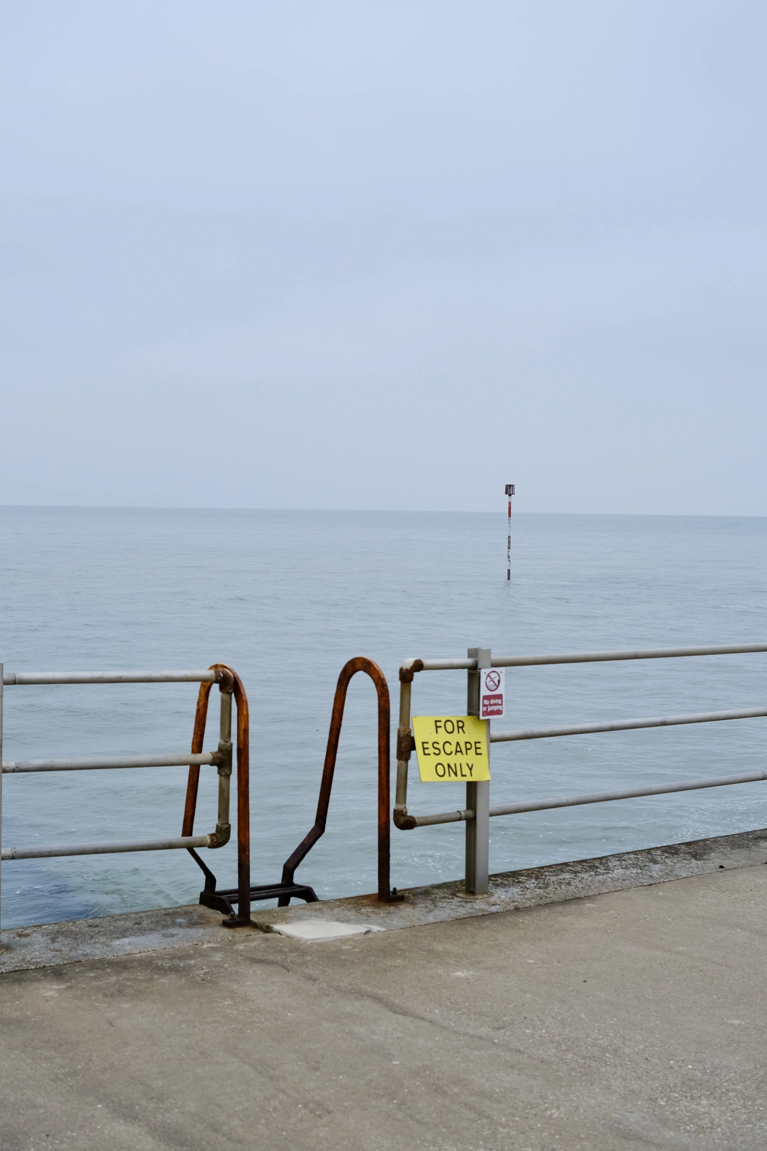 View of the ocean with a metal barrier and a yellow sign that reads 'FOR ESCAPE ONLY'. There is a solitary navigation marker in the water and a cloudy sky above.