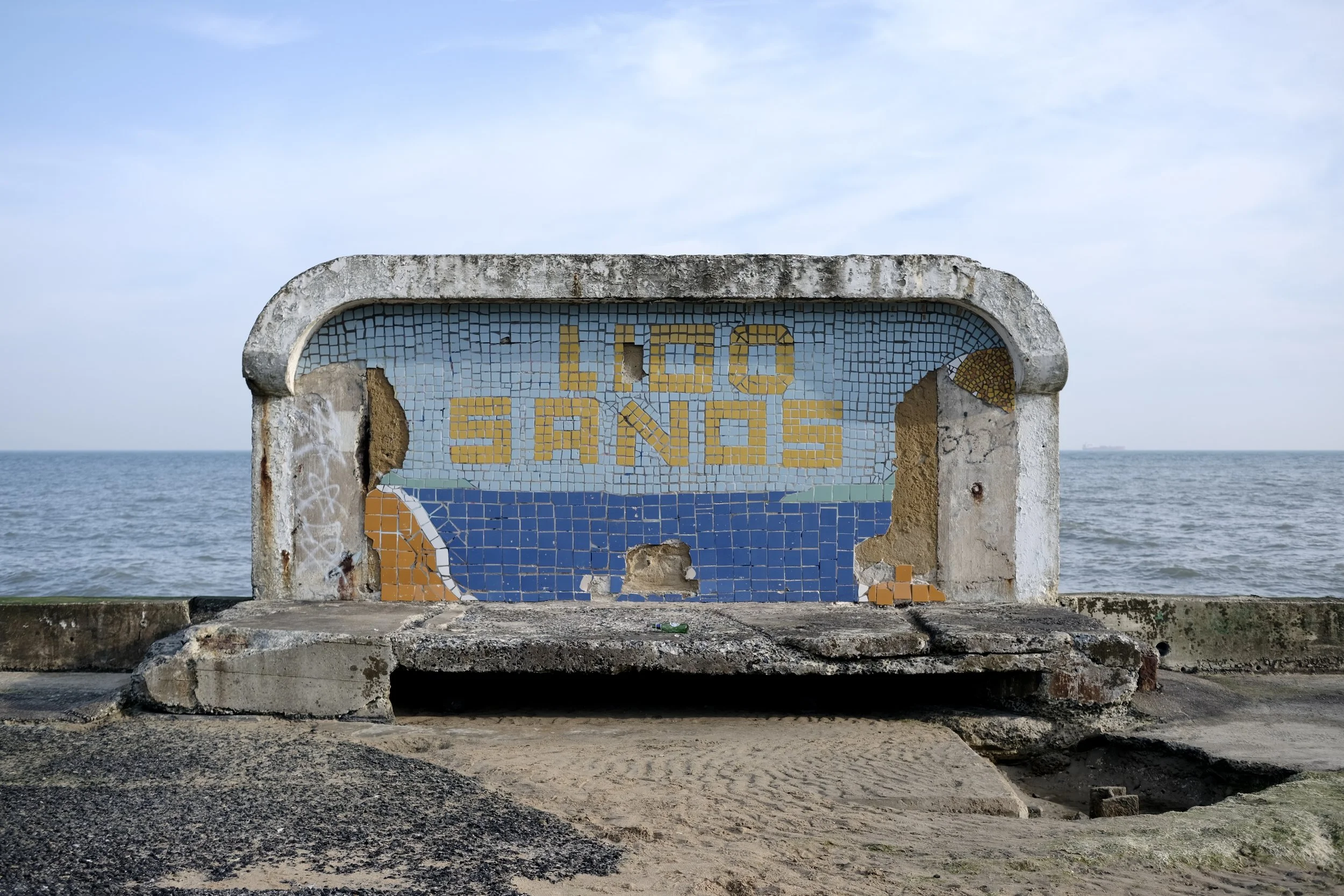A weathered concrete sign with a mosaic design that reads 'LOO SANDOS' in yellow and blue tiles, situated near the beach with the ocean and sky in the background.