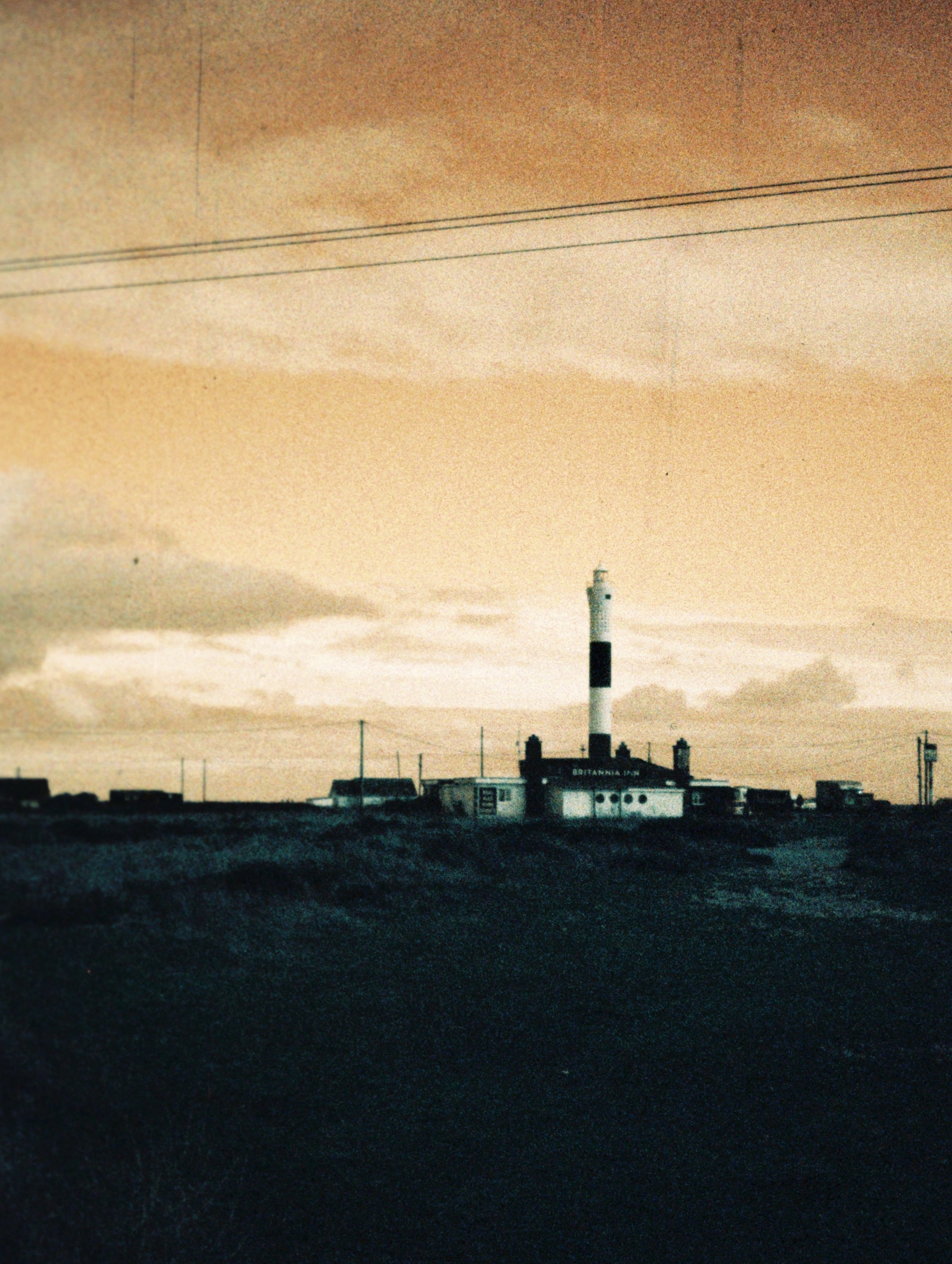 A lighthouse on a coastal landscape during sunset or sunrise, with buildings and power lines in the foreground.