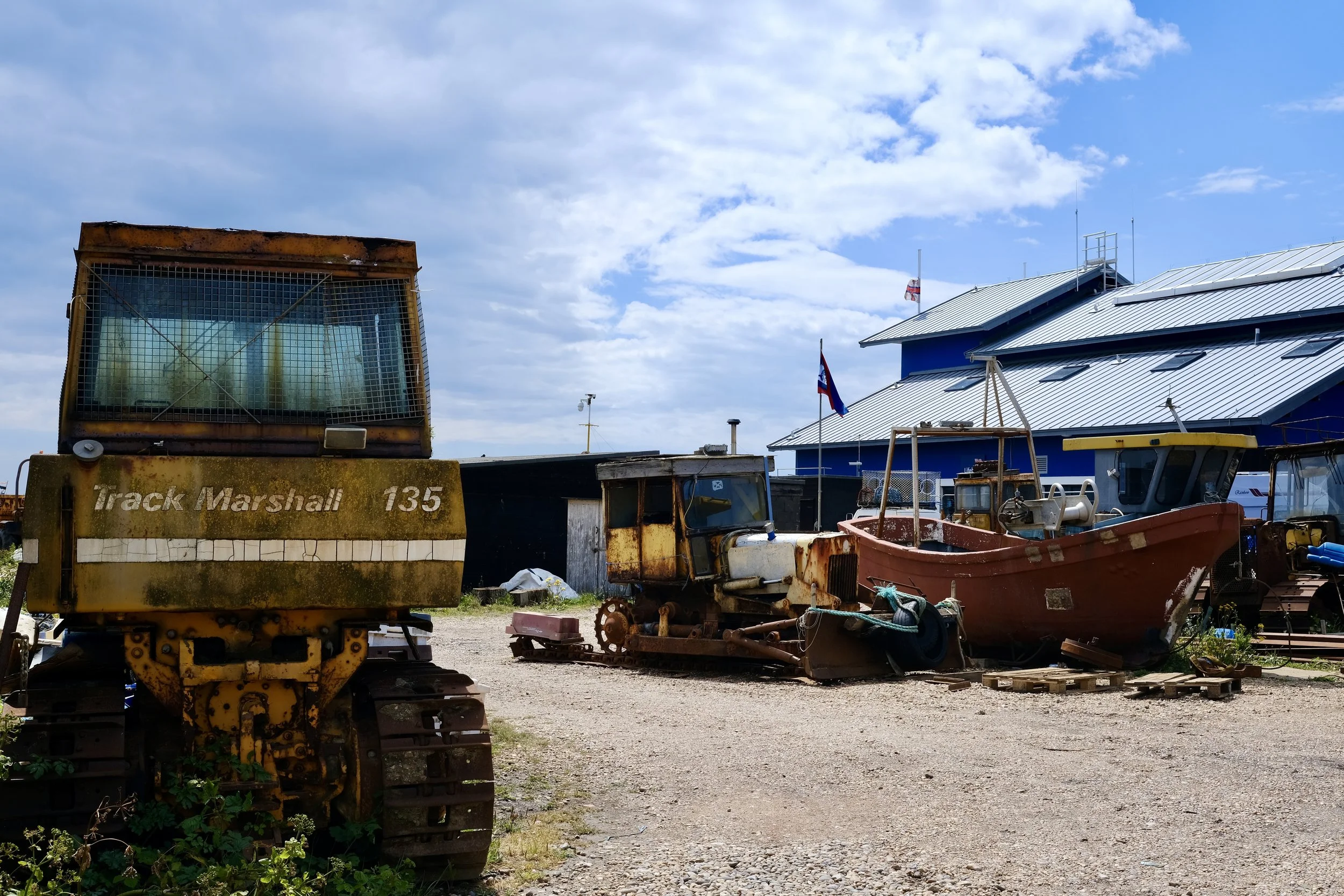 A rusty tracked machine in front of a building with a metal roof, flags, and various boats and equipment in a yard under a partly cloudy sky.