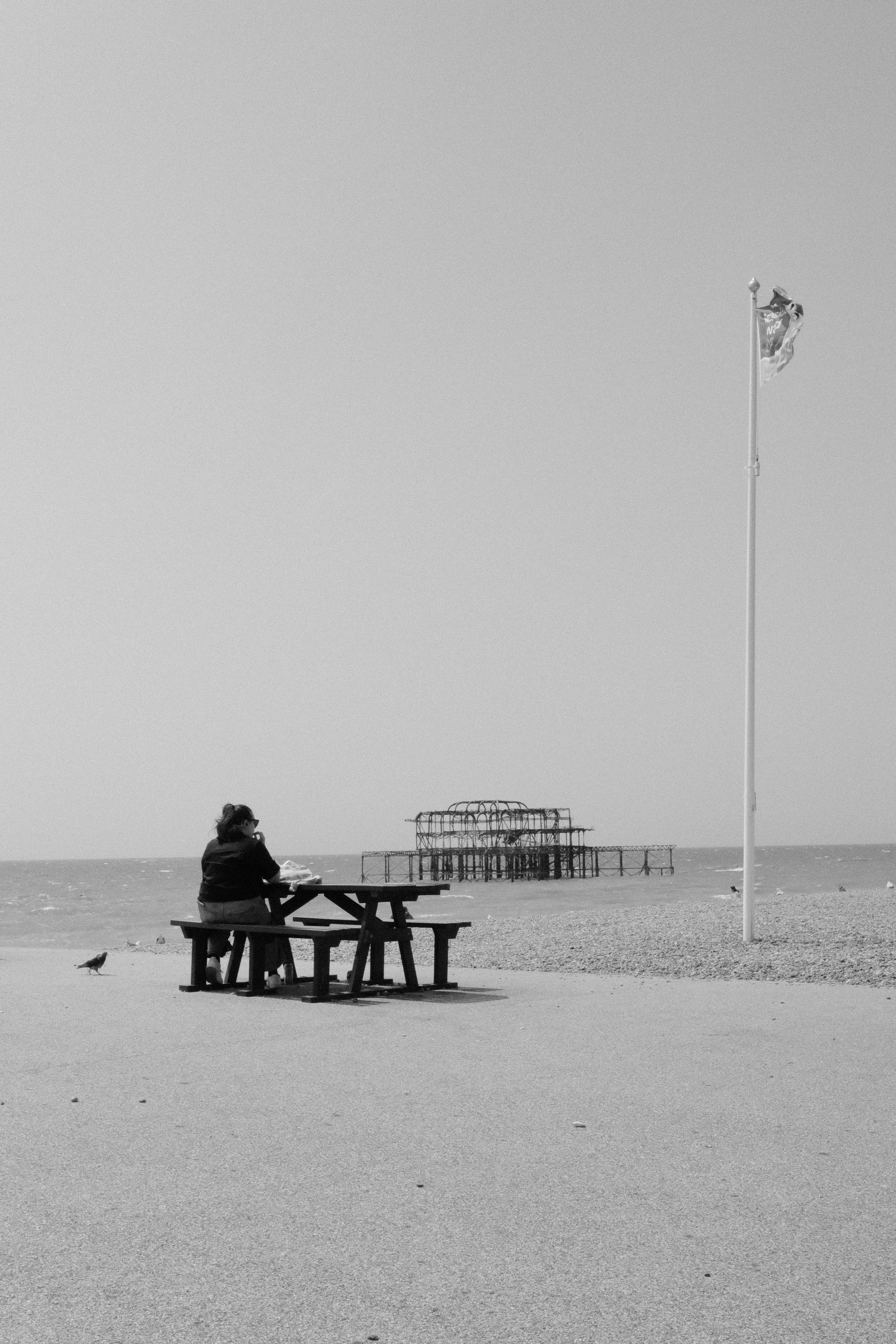 A woman sitting alone at a picnic table on a beach, with a structure in the water in the background and a flagpole with a flag on it.