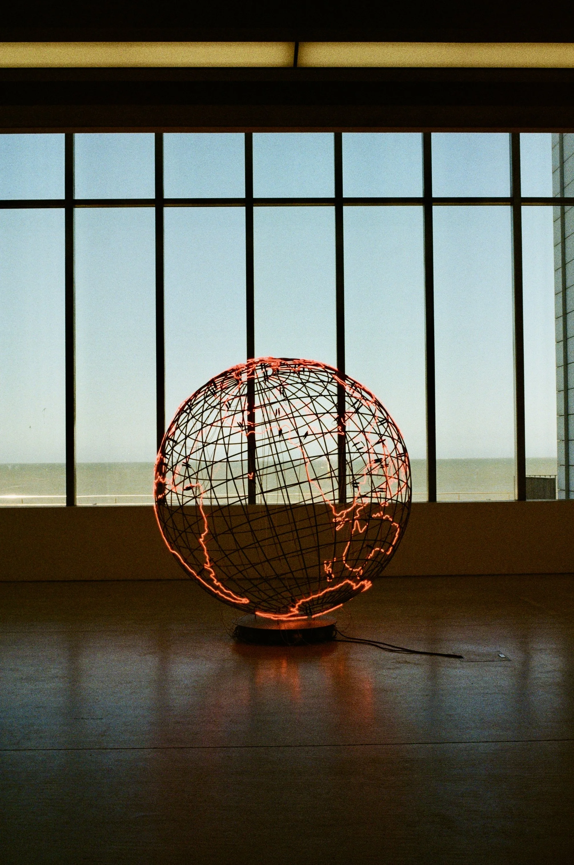A wire globe sculpture of the Earth illuminated with red lights, positioned indoors near a large window showing a blue sky.