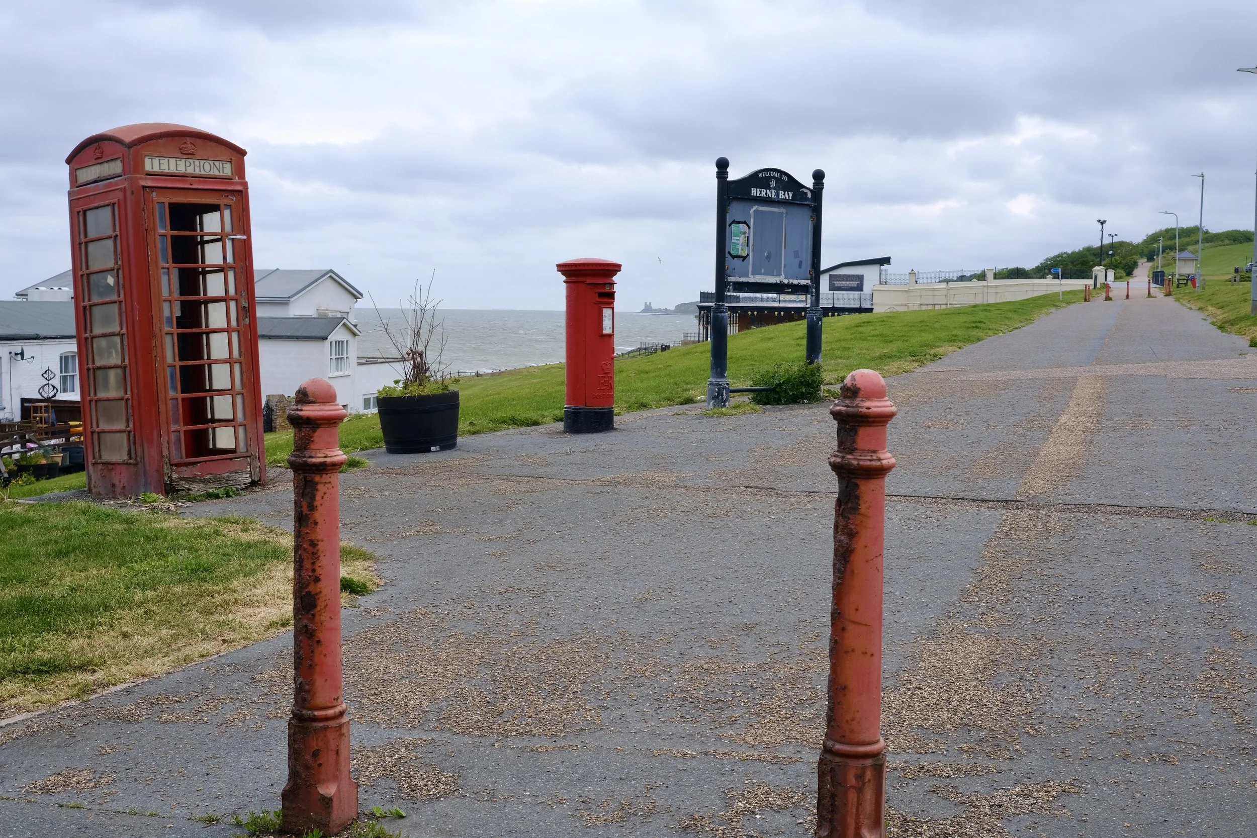Red telephone booth, red post box, black signpost, pathway along coast with grassy area, white buildings, cloudy sky, ocean in background, distant castle on water.