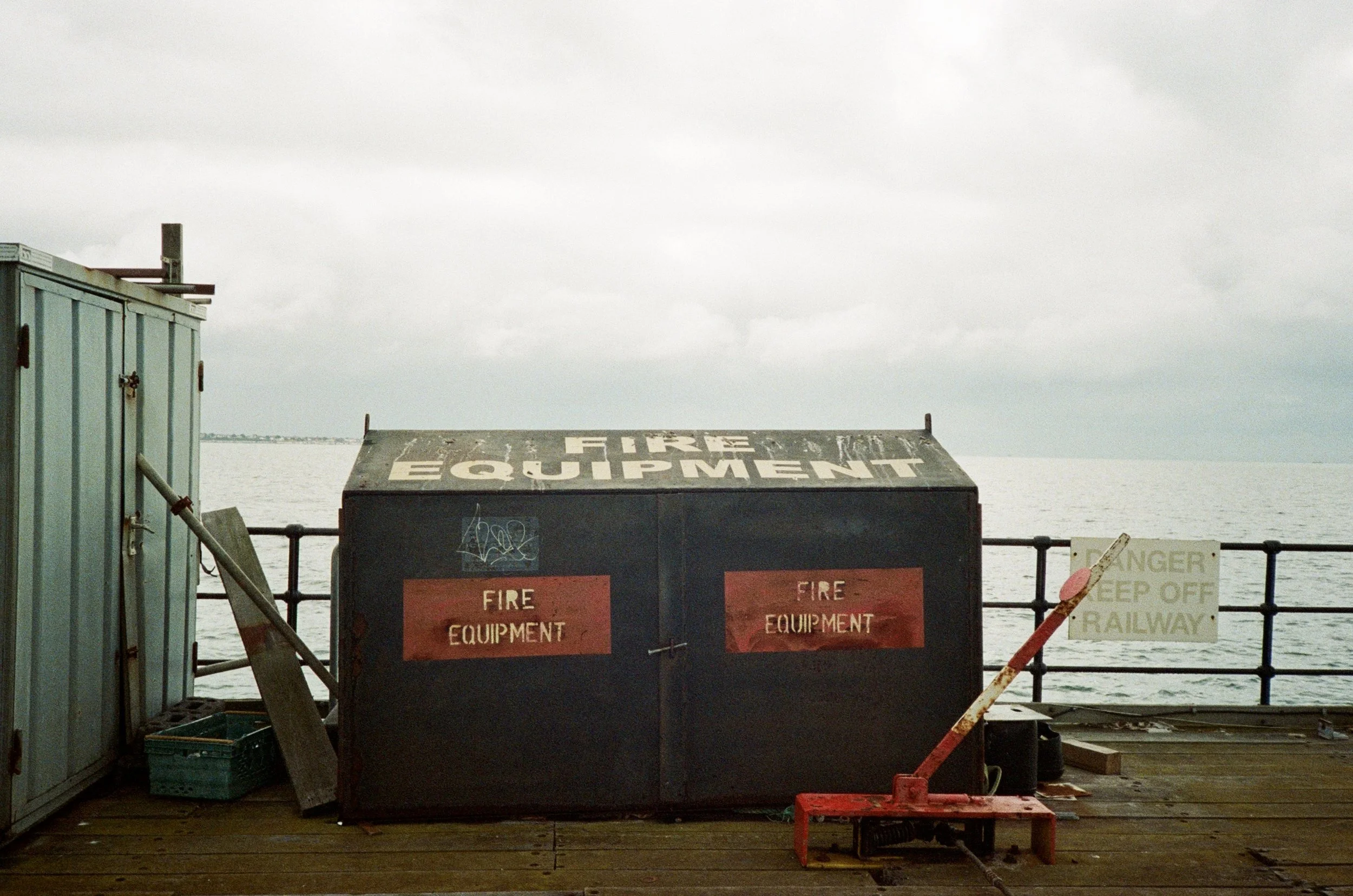 A large black fire equipment cabinet with red warning signs, located outdoors on a wooden deck near the water, with a blue electrical box on the left and some signage on the right, under a cloudy sky.