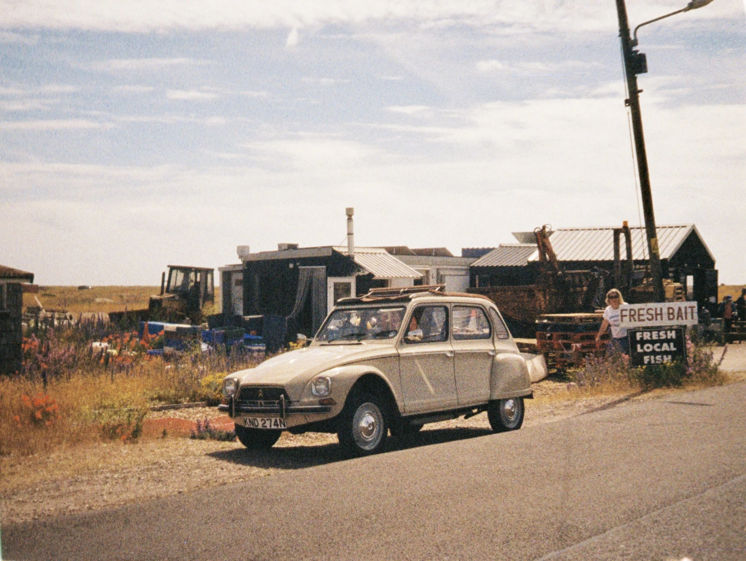Vintage beige car parked beside a sign that reads 'Fresh Bait, Fresh Local Fish' at an outdoor coastal area with buildings and a clear sky.