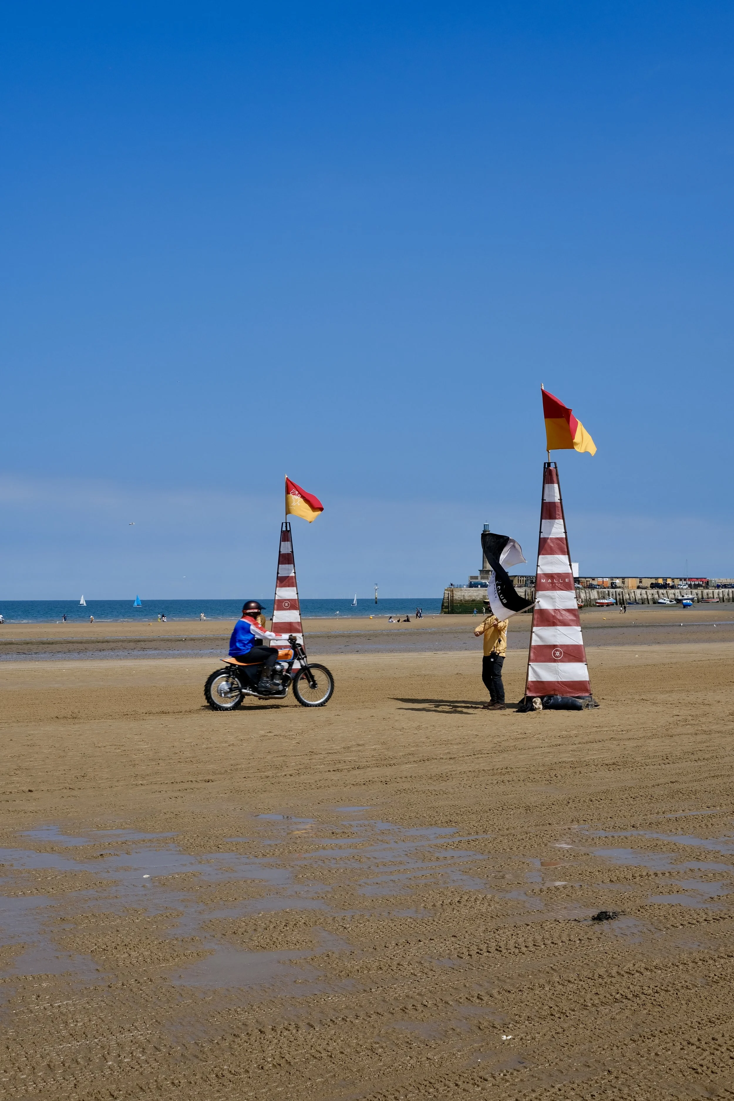A sandy beach with a person riding a motorcycle, two flagpoles with red and yellow flags, and a person standing near the flagpoles. In the background, the shoreline with sailboats, a building, and a clear blue sky.