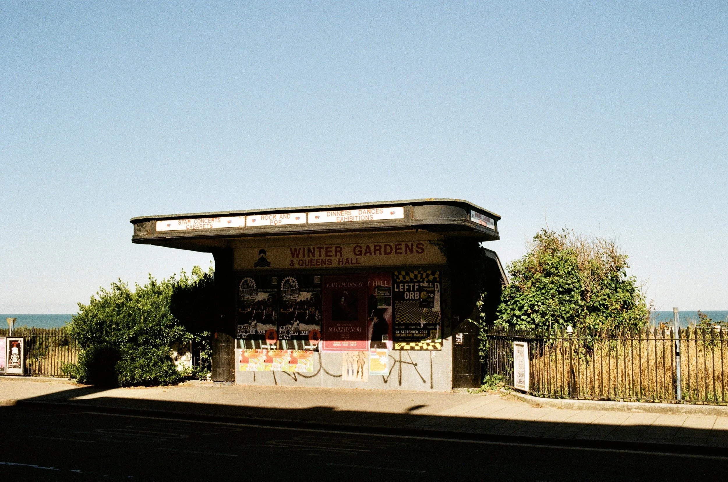 A small outdoor structure advertising events such as star concerts, cabaret acts, rock and pop music, dinners, dances, and exhibitions, with trees and a clear sky in the background.