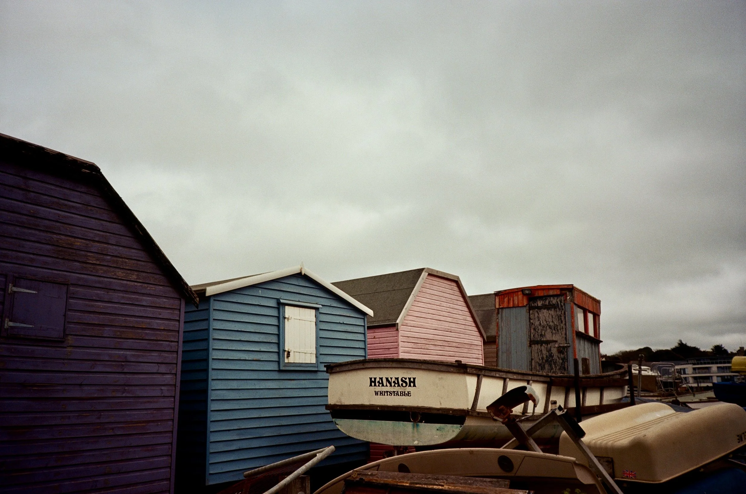 Colorful beach huts on a cloudy day, with a boat named 'HANASH' in the foreground.