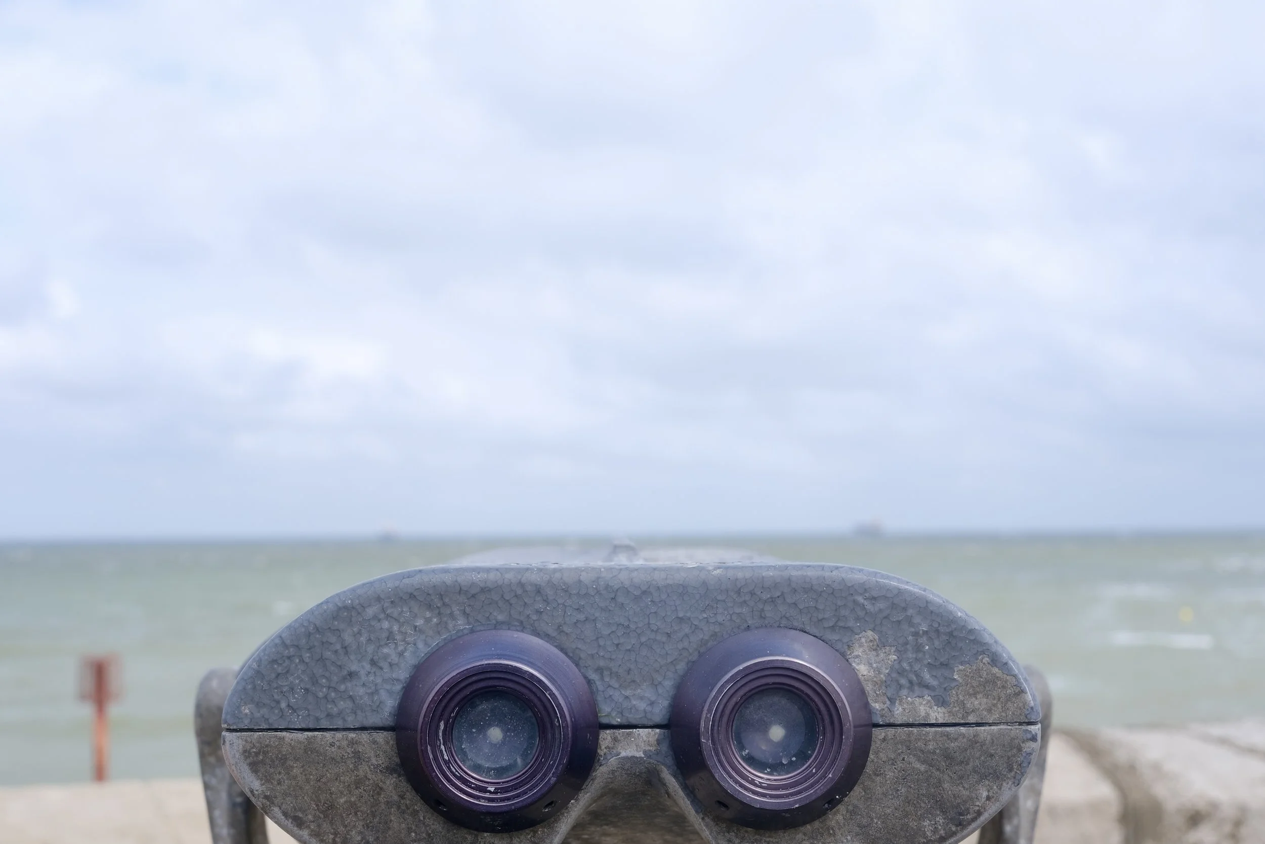 A view through a pair of binoculars on a tripod, looking out at a cloudy sky over a body of water in the distance.
