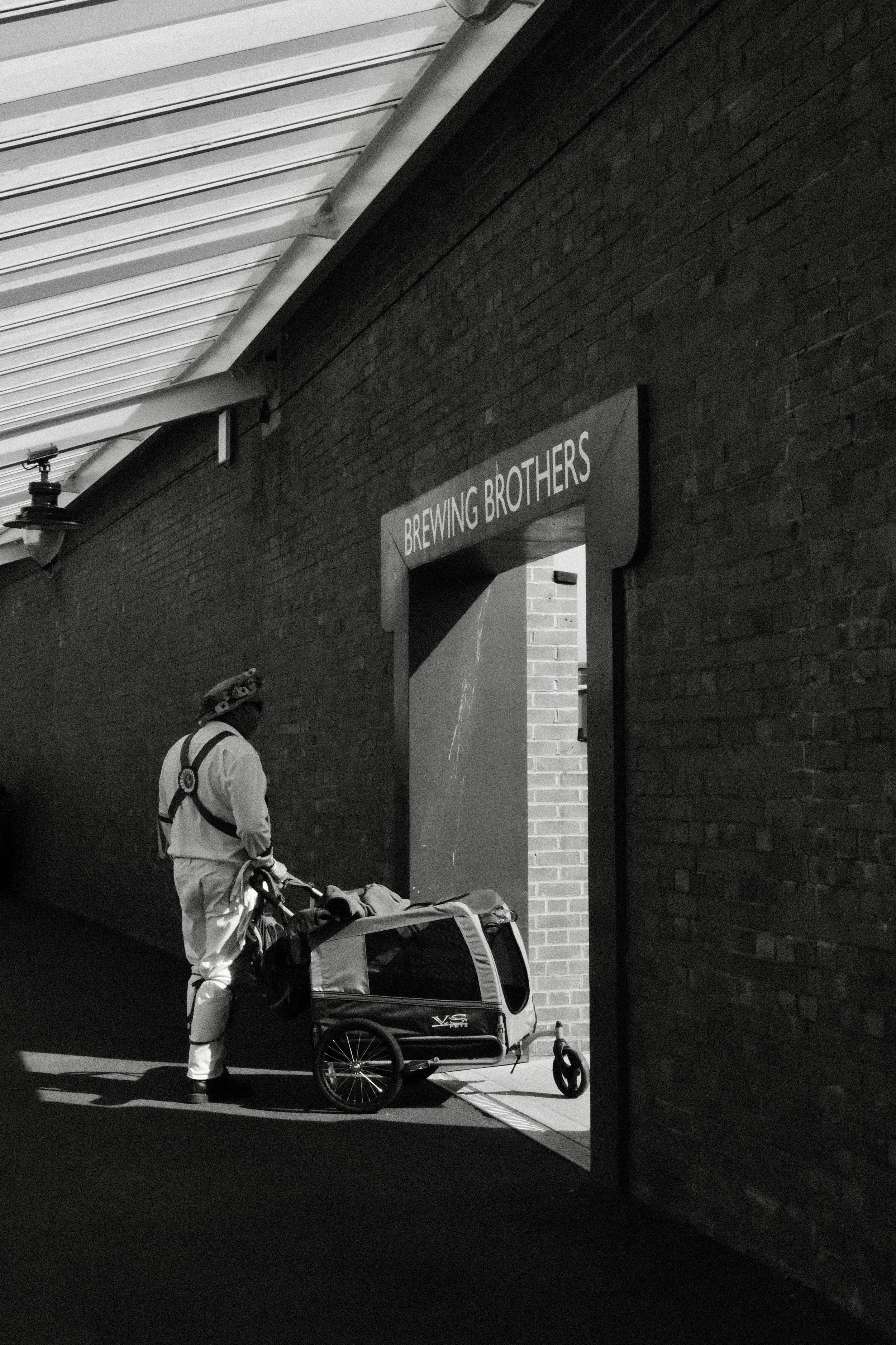 A person in protective gear standing next to a stroller with a pet carrier attached, in an outdoor covered walkway area with a brick wall and a sign reading 'Brewing Brothers'.