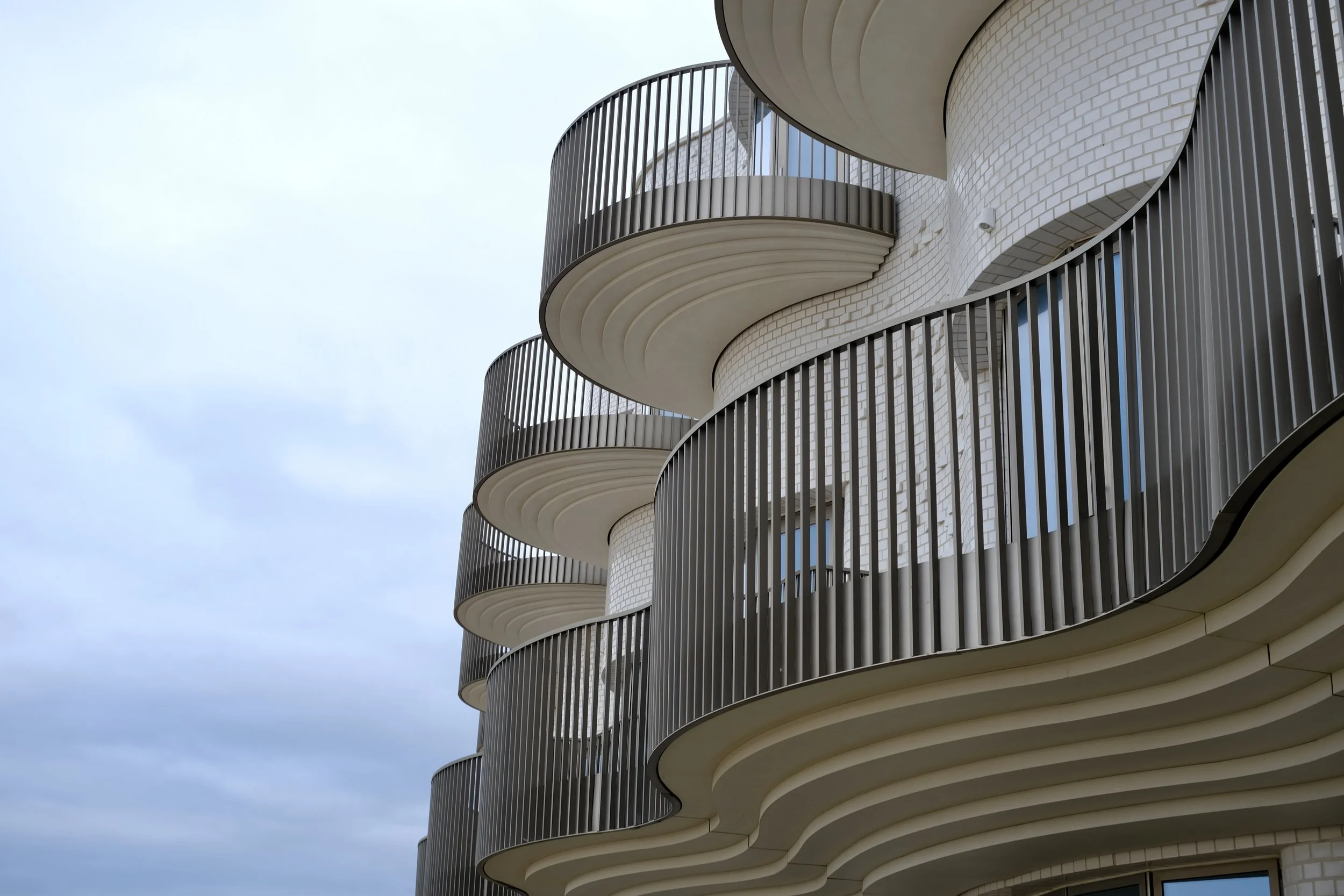 Close-up of a modern building with curved balconies and vertical metal railings against a cloudy sky.