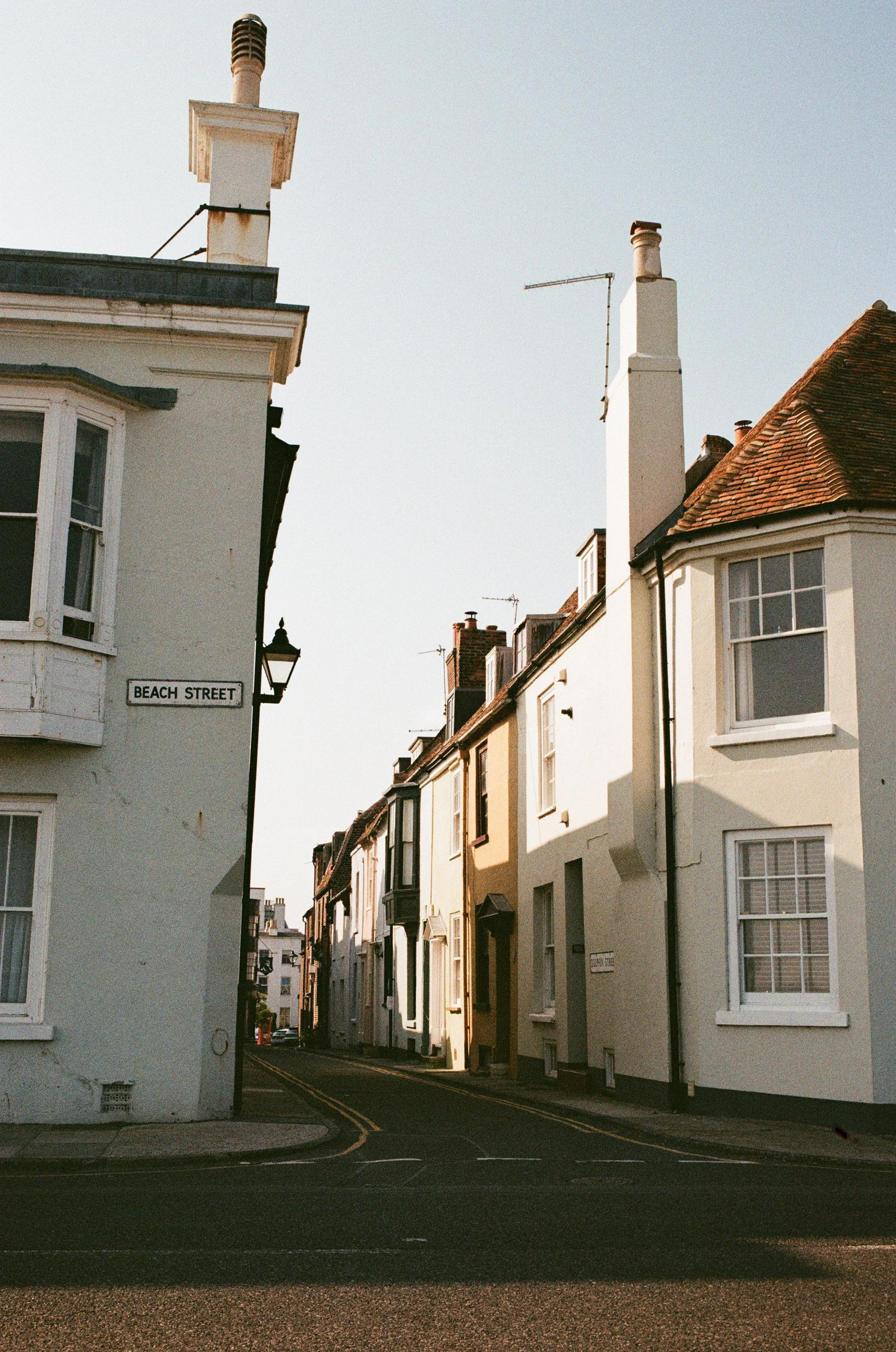 A narrow street lined with white houses, some with bay windows, and a street sign reading 'Beach Street' on the corner of one house. The sky is clear and the lighting suggests late afternoon or early evening.