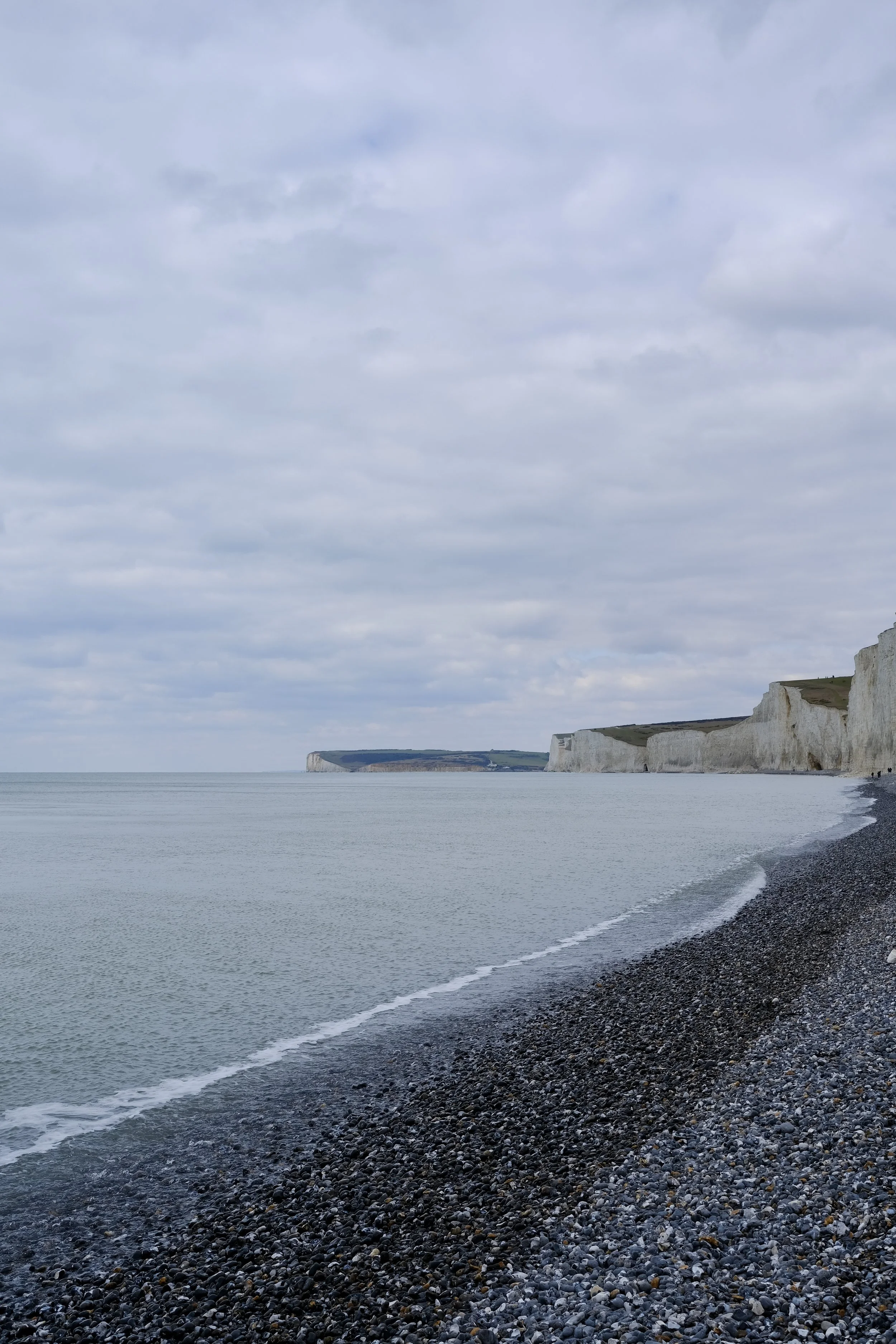 Cliffs overlooking a pebble beach and calm sea with cloudy sky.