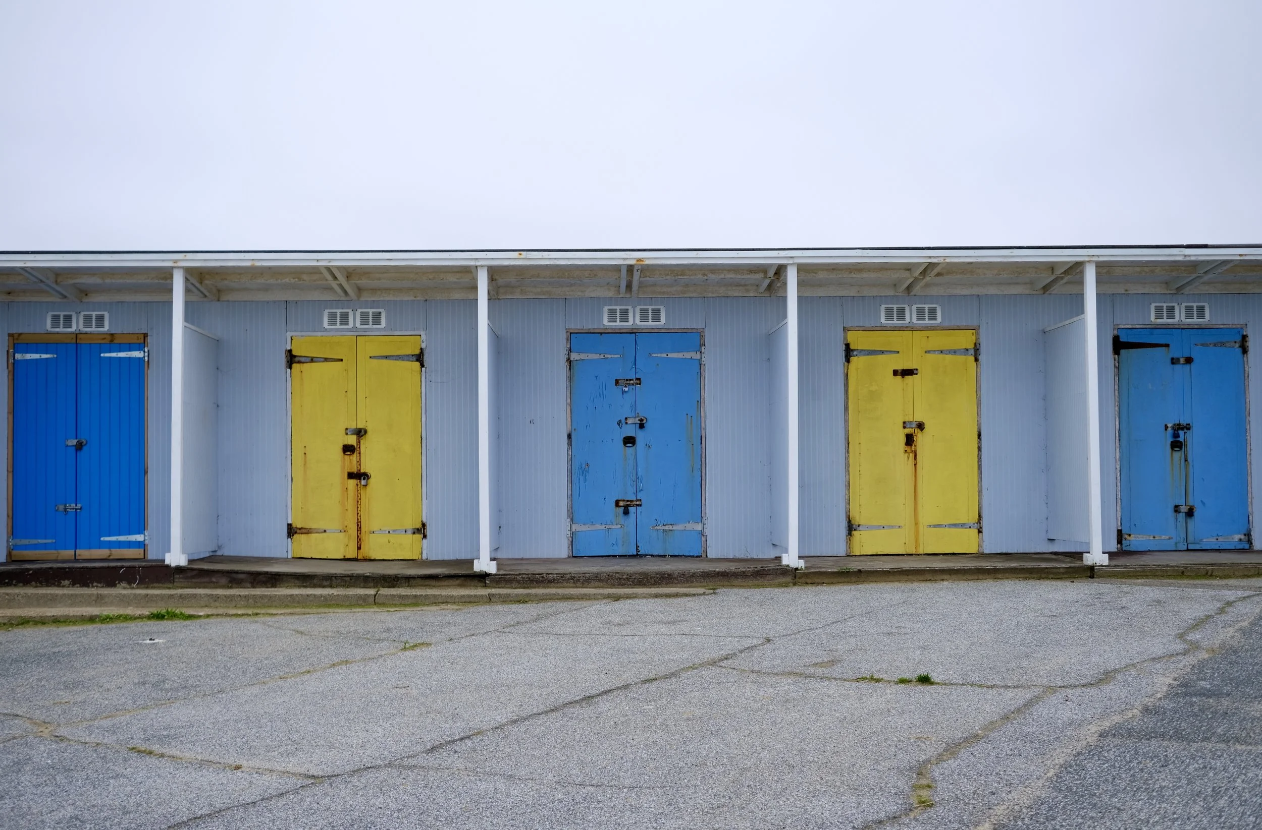 Colorful storage units with yellow and blue doors sit under a coral-colored roof, with an empty parking lot in front and a cloudy sky overhead.