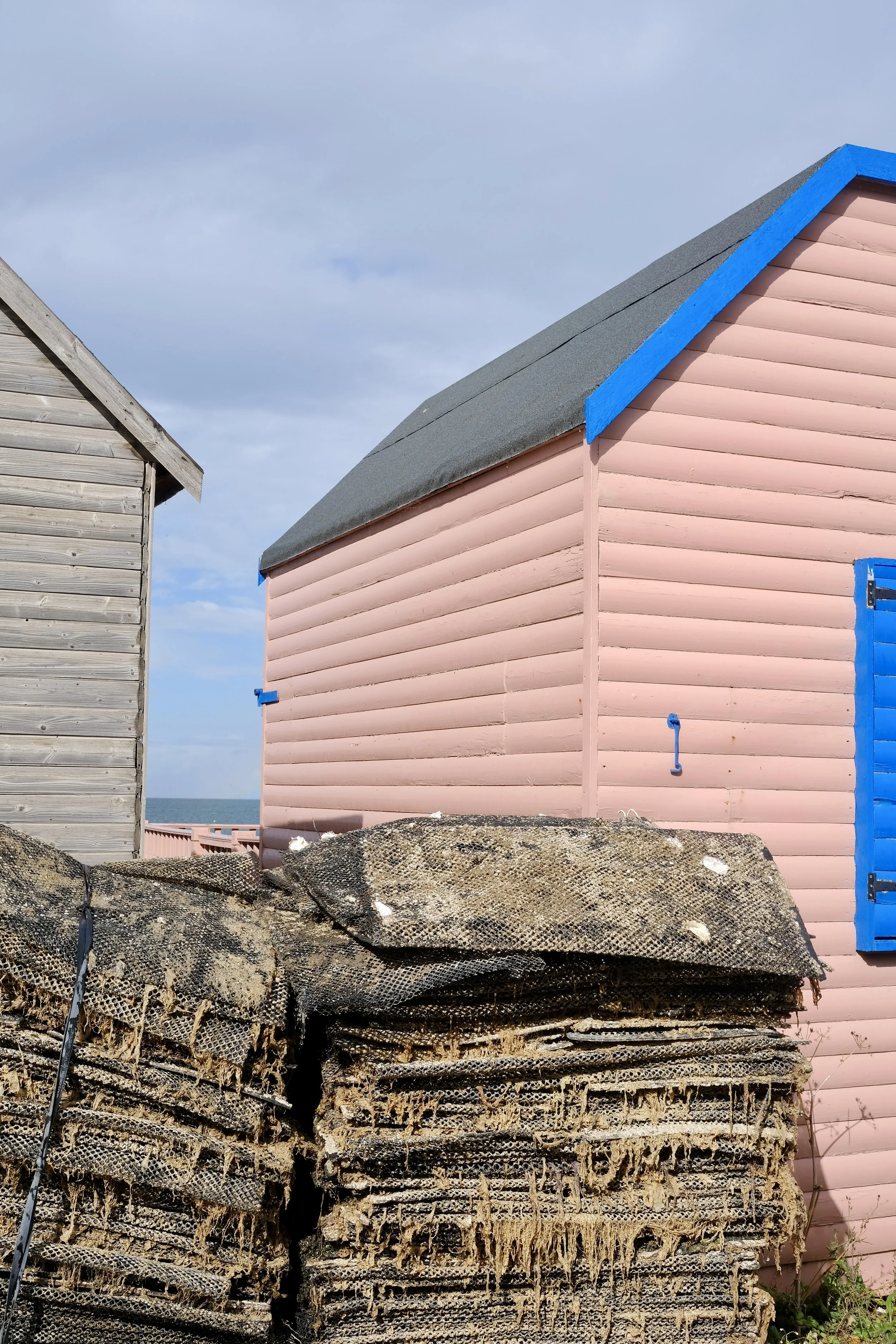 A row of weathered lobster traps in the foreground, with two beach houses in the background, one gray and unpainted, the other pink with blue trim, under a cloudy sky.