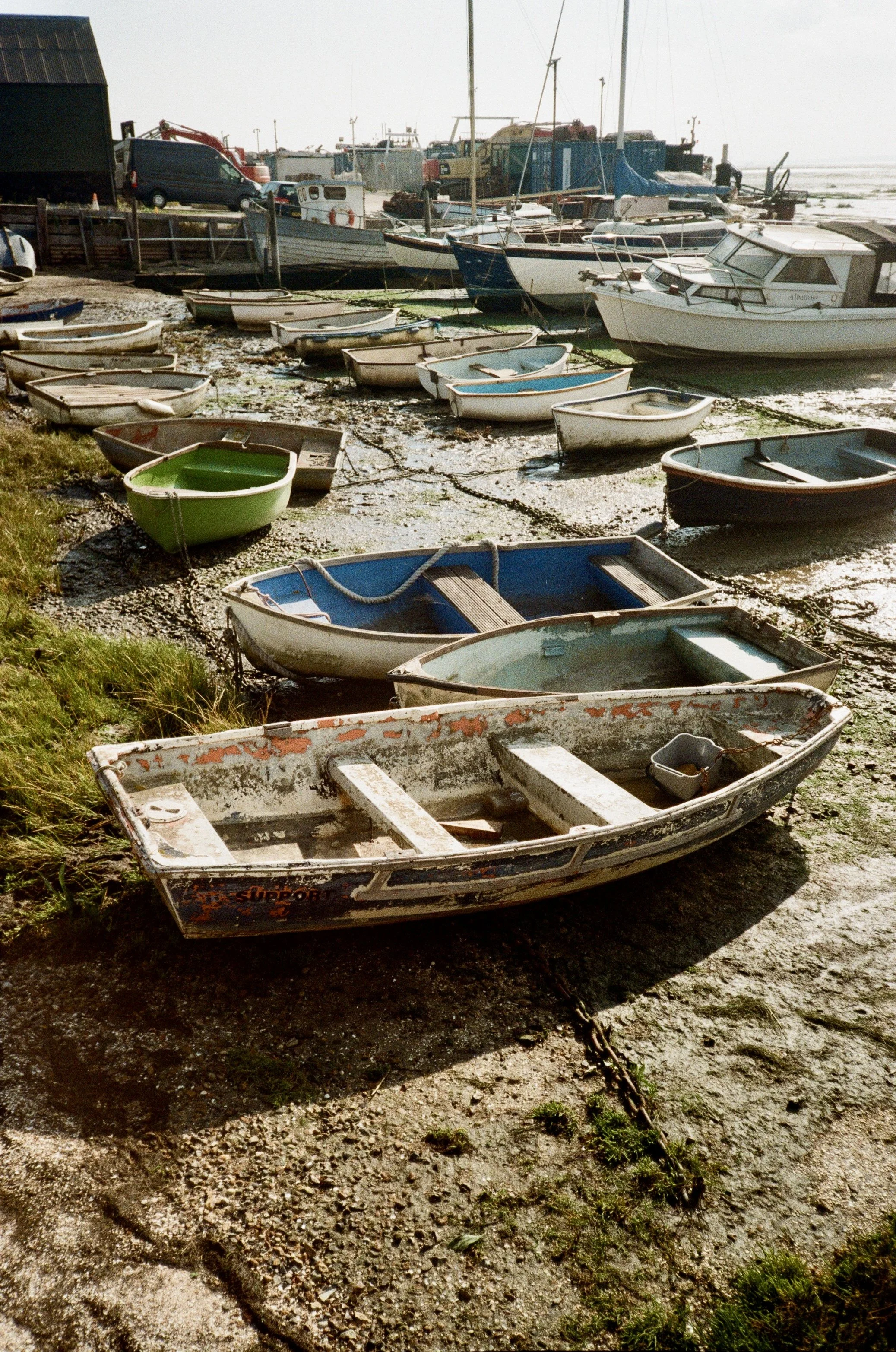 Multiple small boats on muddy ground near water, some with chains, some with hulls showing visible wear, and a large docked boat in the background along with a parking lot and vehicles.