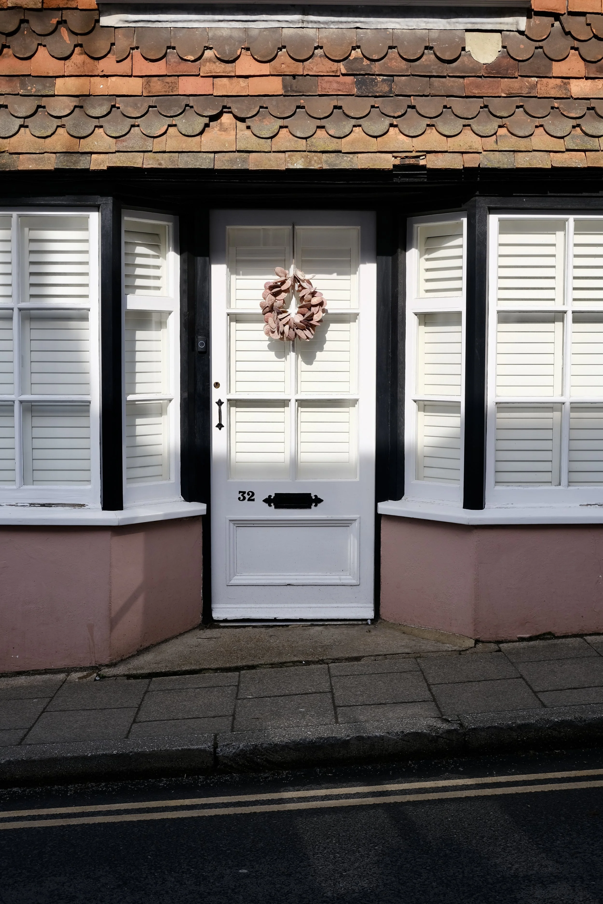 White front door with house number 32 and a black mailbox, flanked by two window shutters with white blinds, and a pink wreath hanging on the door, with a brick-tiled roof above.