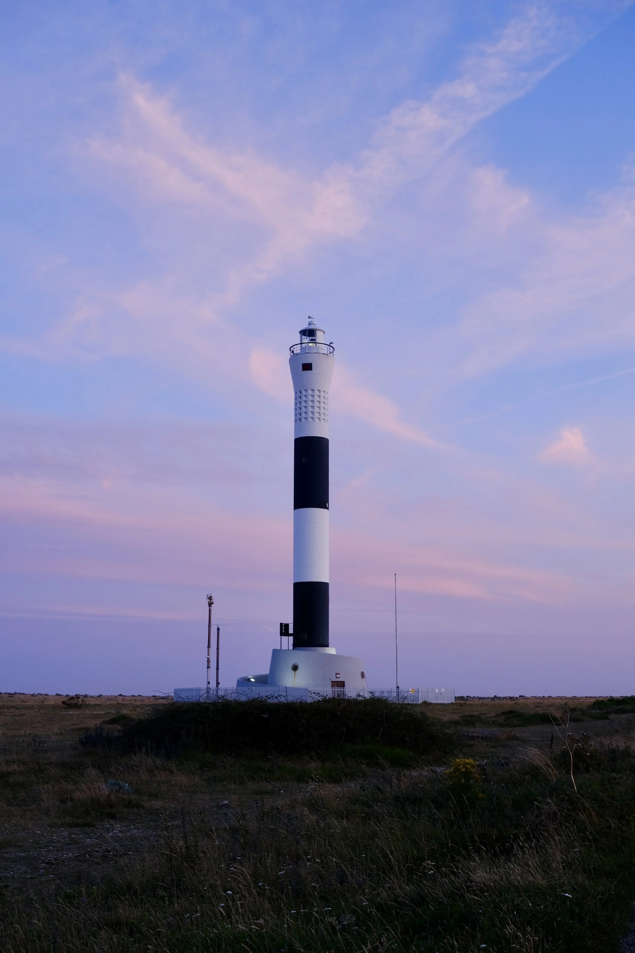 A white lighthouse with black stripes against a sky with pink and blue clouds, surrounded by grass and a fence.