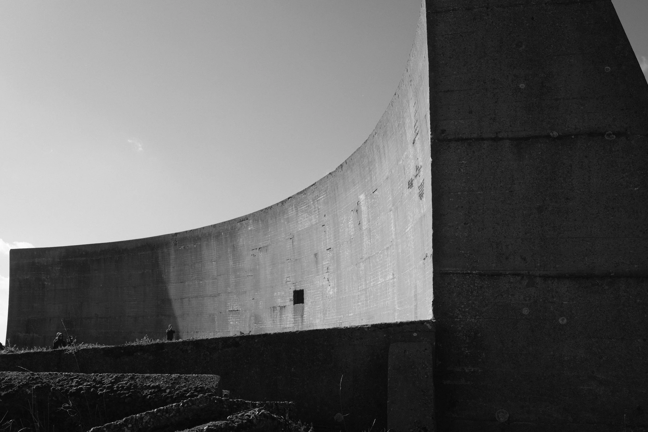 A black and white photo of a large concrete structure, possibly a dam, with people standing at its base and the sky in the background.