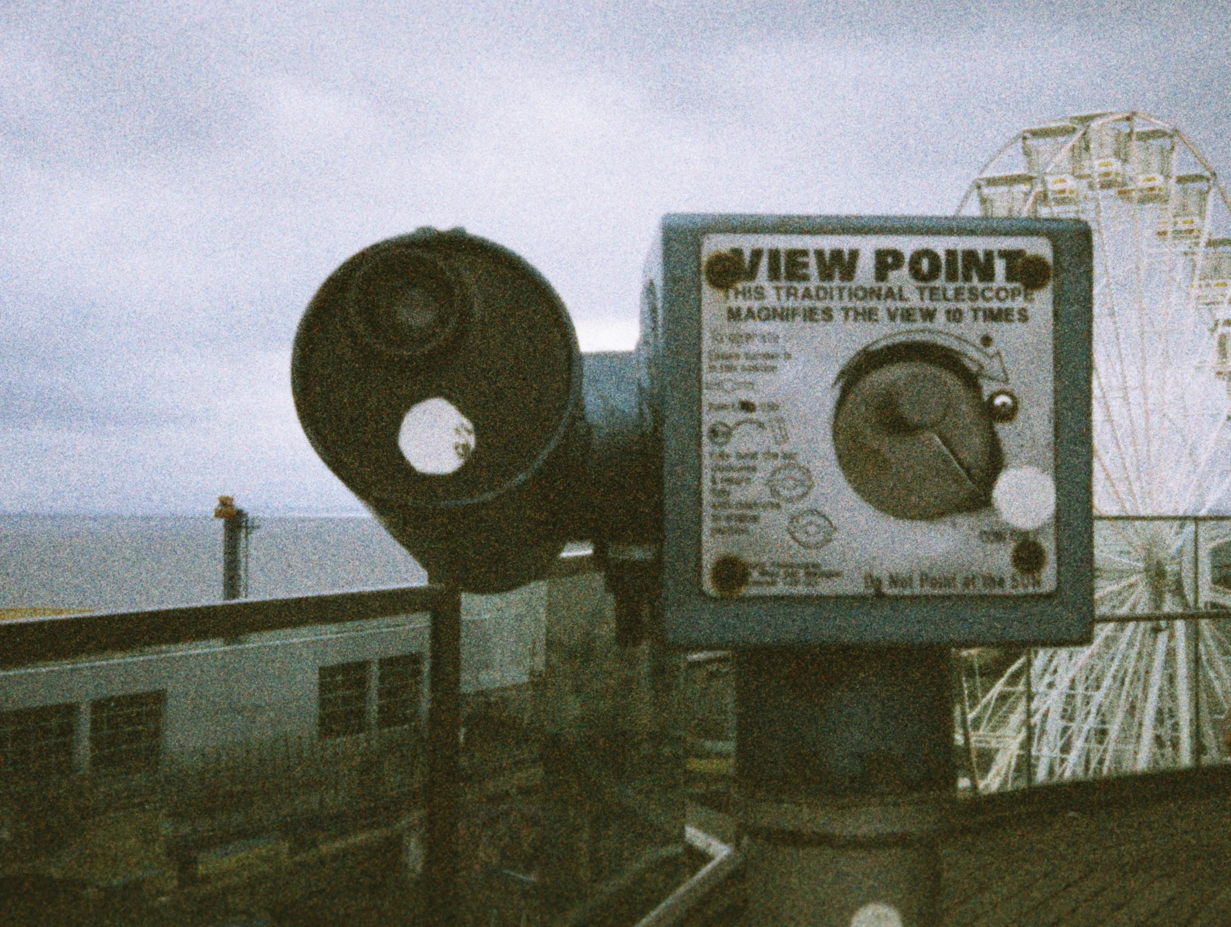 The image shows a signboard for View Point with a description about a traditional telescope that magnifies the view to tens of miles. In the background, there is a Ferris wheel and some industrial structures. The atmosphere appears cloudy and the pho