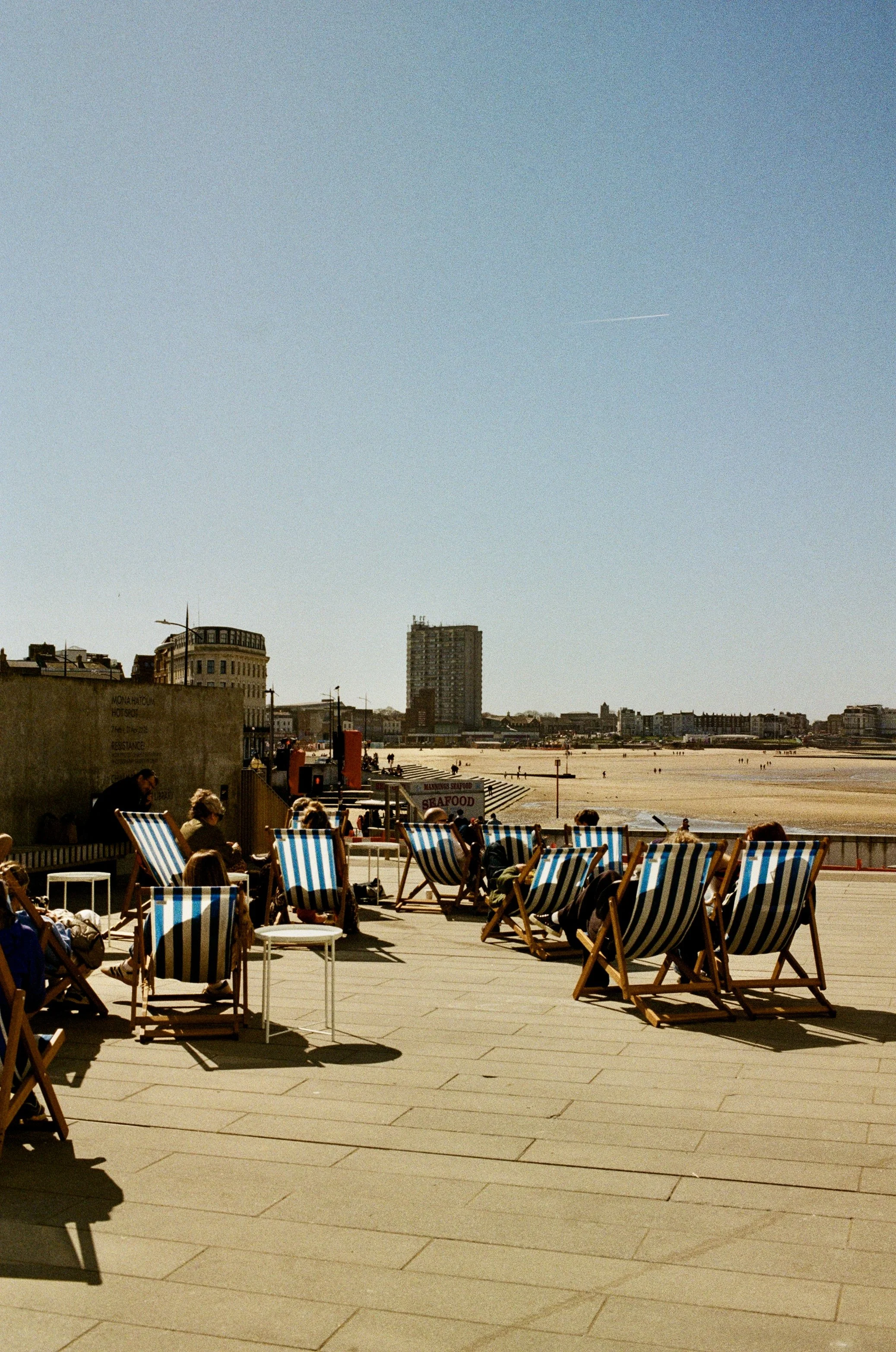 People sitting and relaxing on striped deck chairs on a boardwalk near the beach, with city buildings and a sandy beach in the background under a clear blue sky.