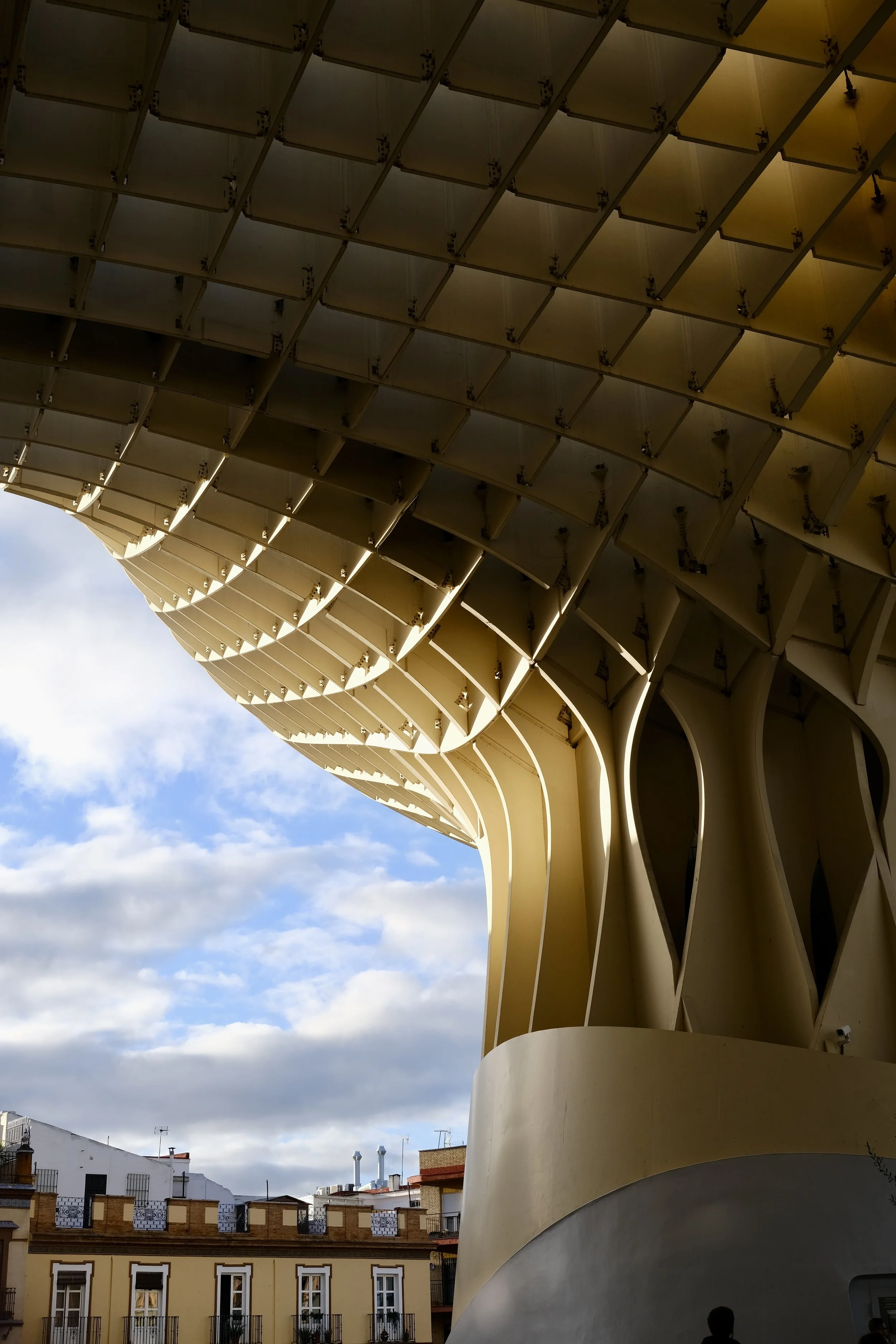 Close-up of the exterior architecture of the C. N. A. (Contemporary Art building), featuring beige curved panels and a metal grid structure, with a cloudy sky in the background.