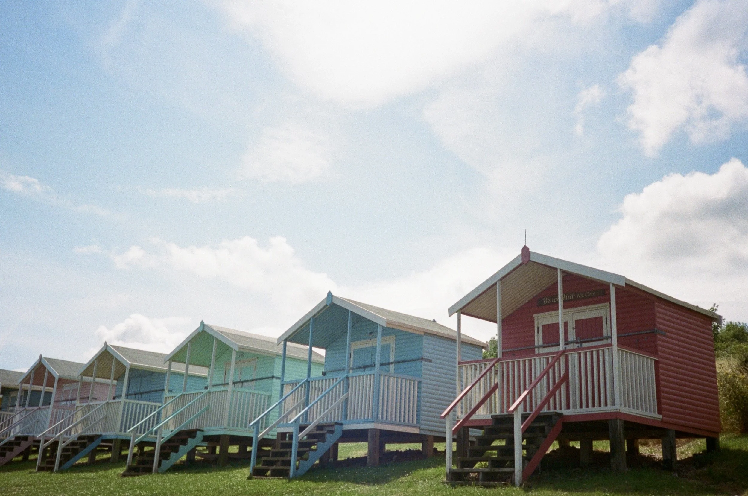Colorful beach huts on stilts with stairs leading up to their entrances, set against a partly cloudy sky.