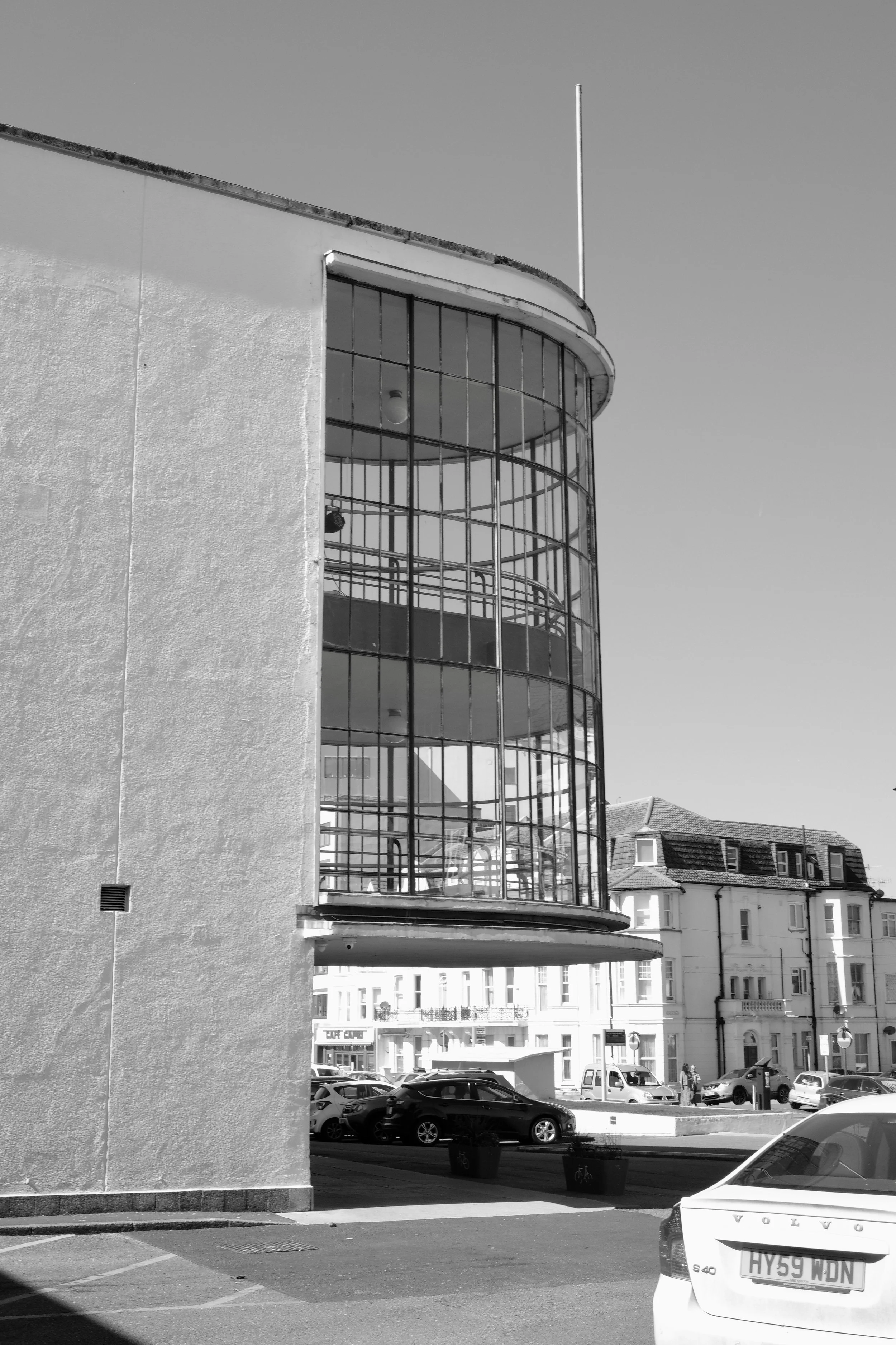 A black and white photo of a city street with cars parked along the side and a modern building with large glass windows and curved architecture.