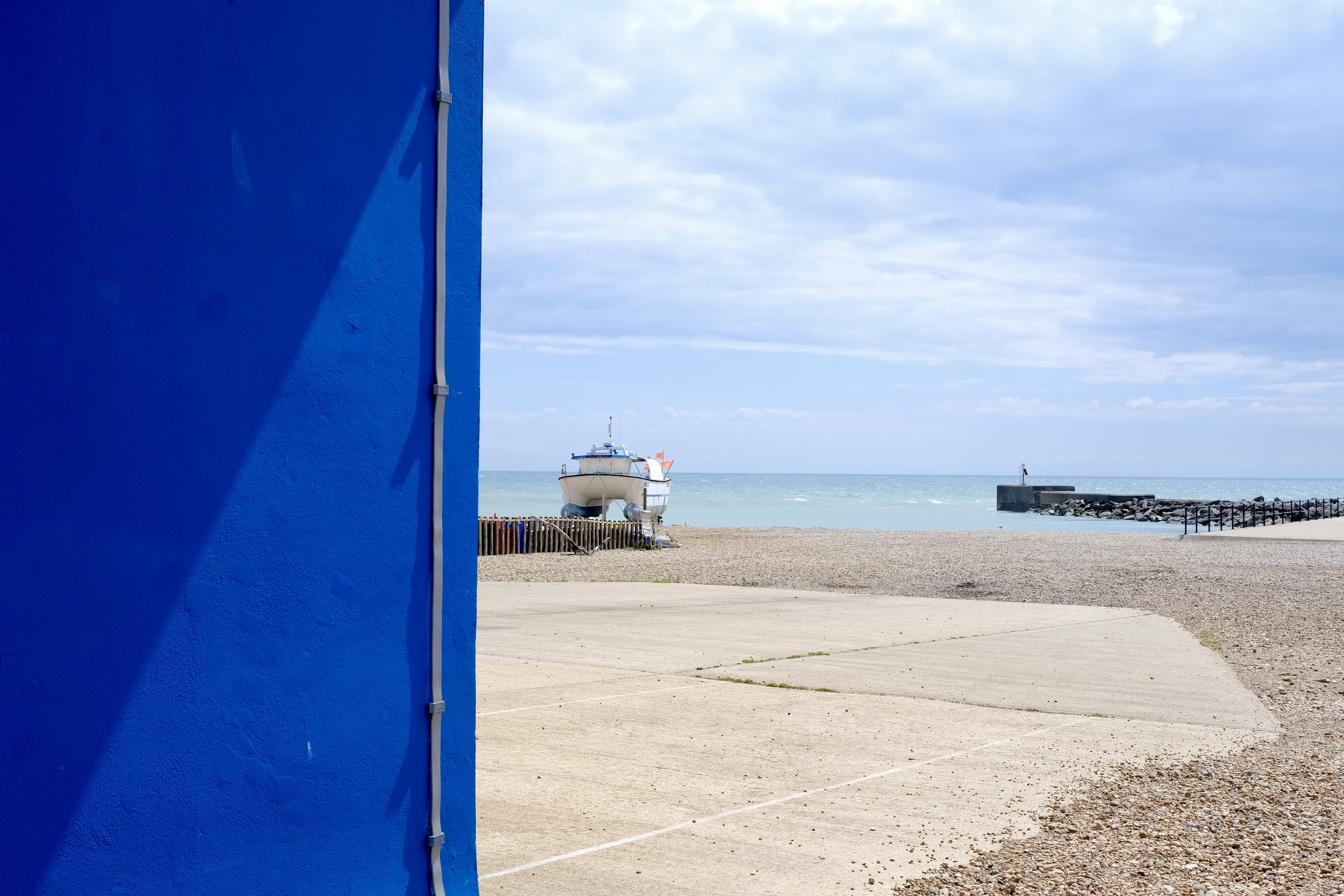 A blue building wall with visible pipes, a sandy pathway, a boat on the water, and cloudy sky.
