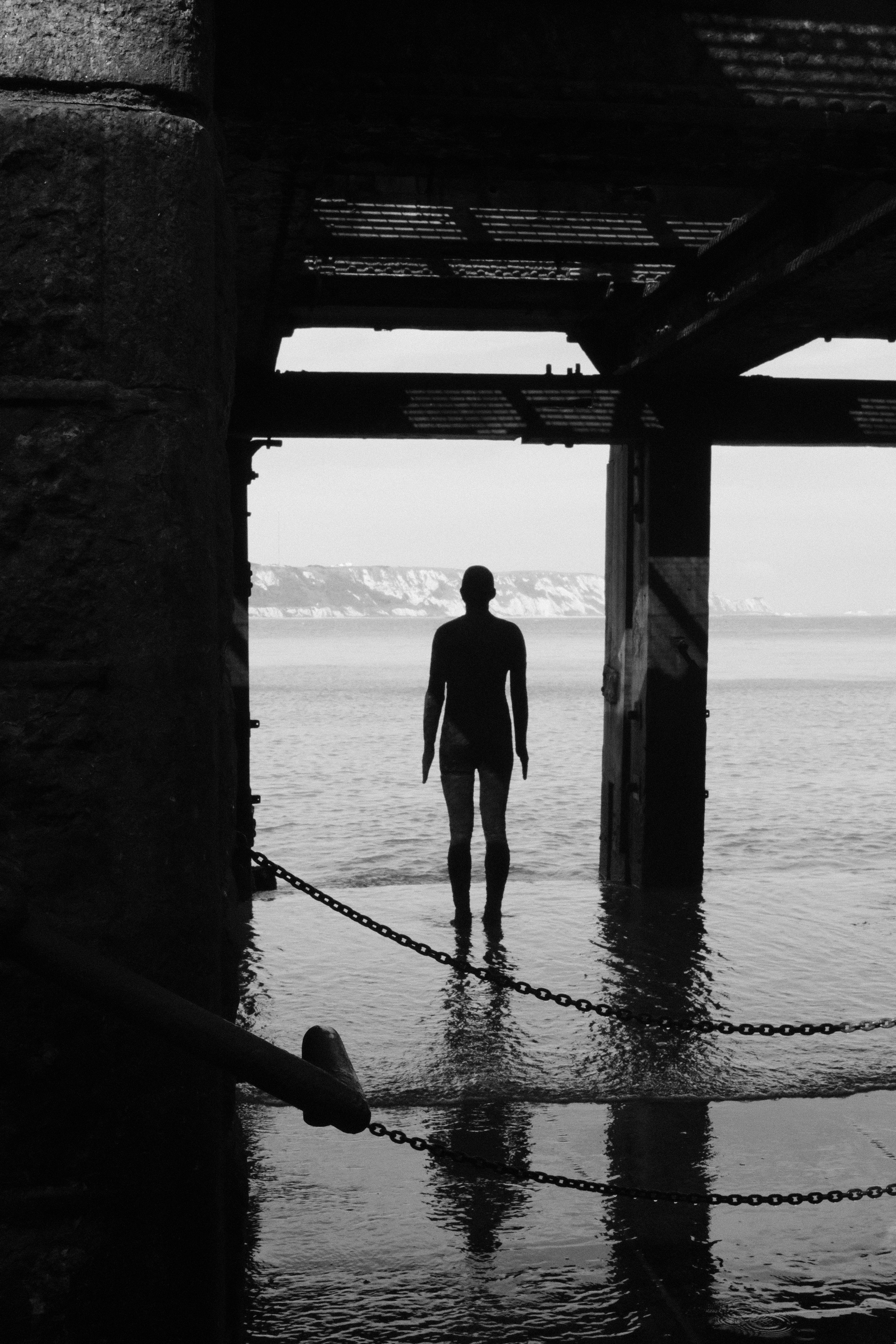 A person standing in water beneath a wooden pier or jetty, with cliffs in the distance, seen through an opening in a stone structure or wall, in black and white.