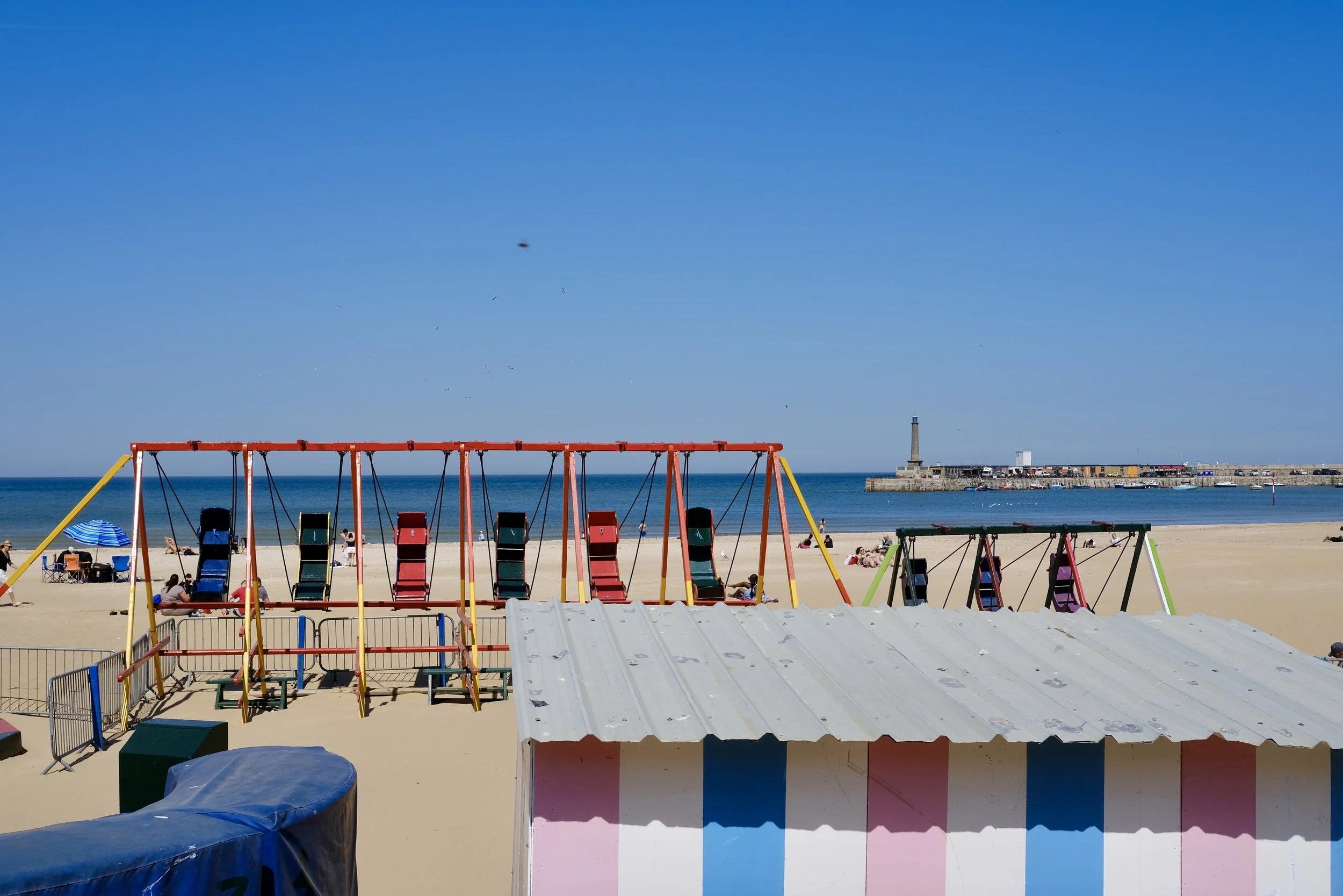 Colorful swings on a sandy beach with a lighthouse and building in the background under a clear blue sky.