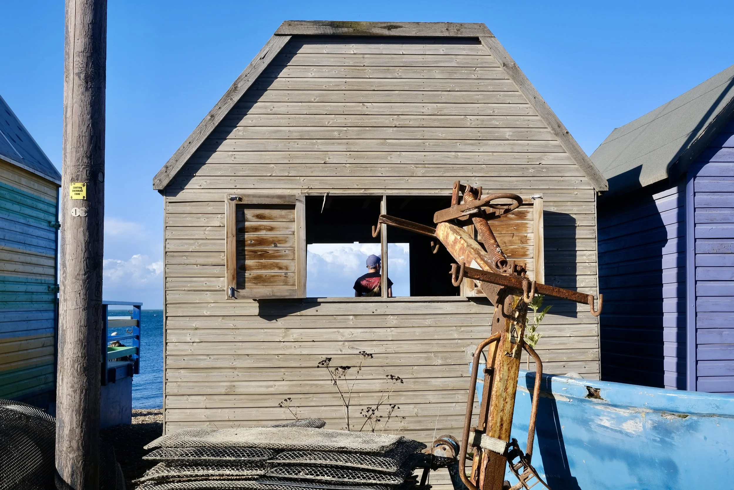 A weathered wooden building under construction with two open window frames, surrounded by colorful beach houses and blue sky, with a rusted anchor sculpture in the foreground.