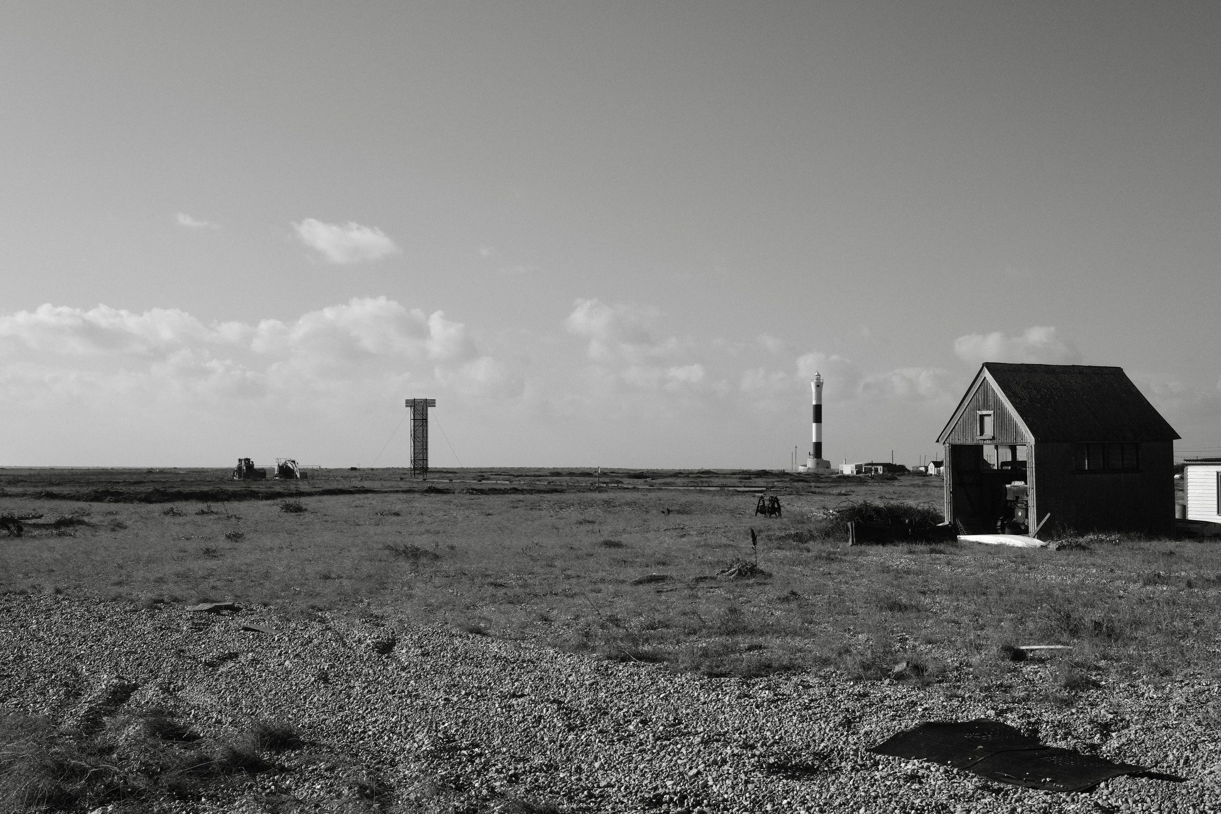 A black and white landscape with a small house on the right, a lighthouse in the distance, and a tall, narrow structure in the center, all under a cloudy sky.