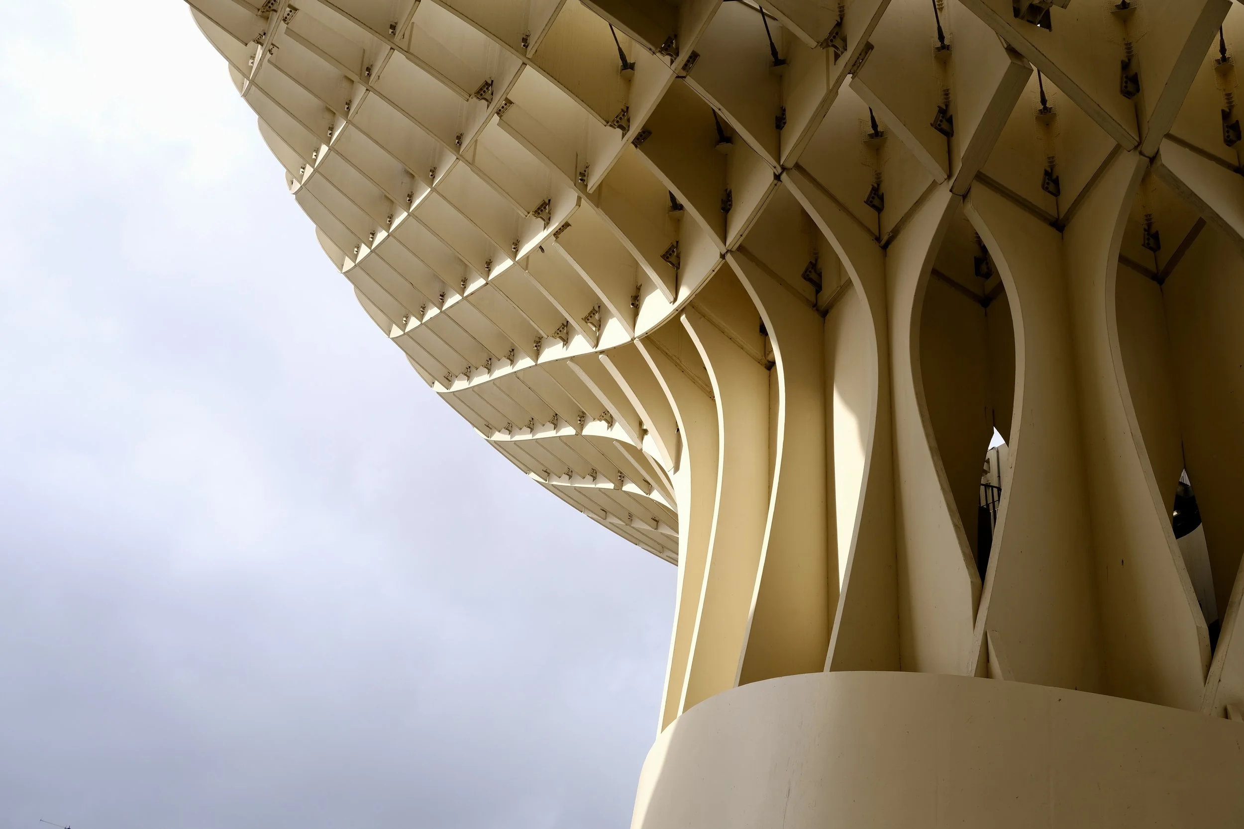 Close-up of a modern architectural structure with a series of yellow wavy panels and supporting beams, viewed from below against a light blue sky with some clouds.