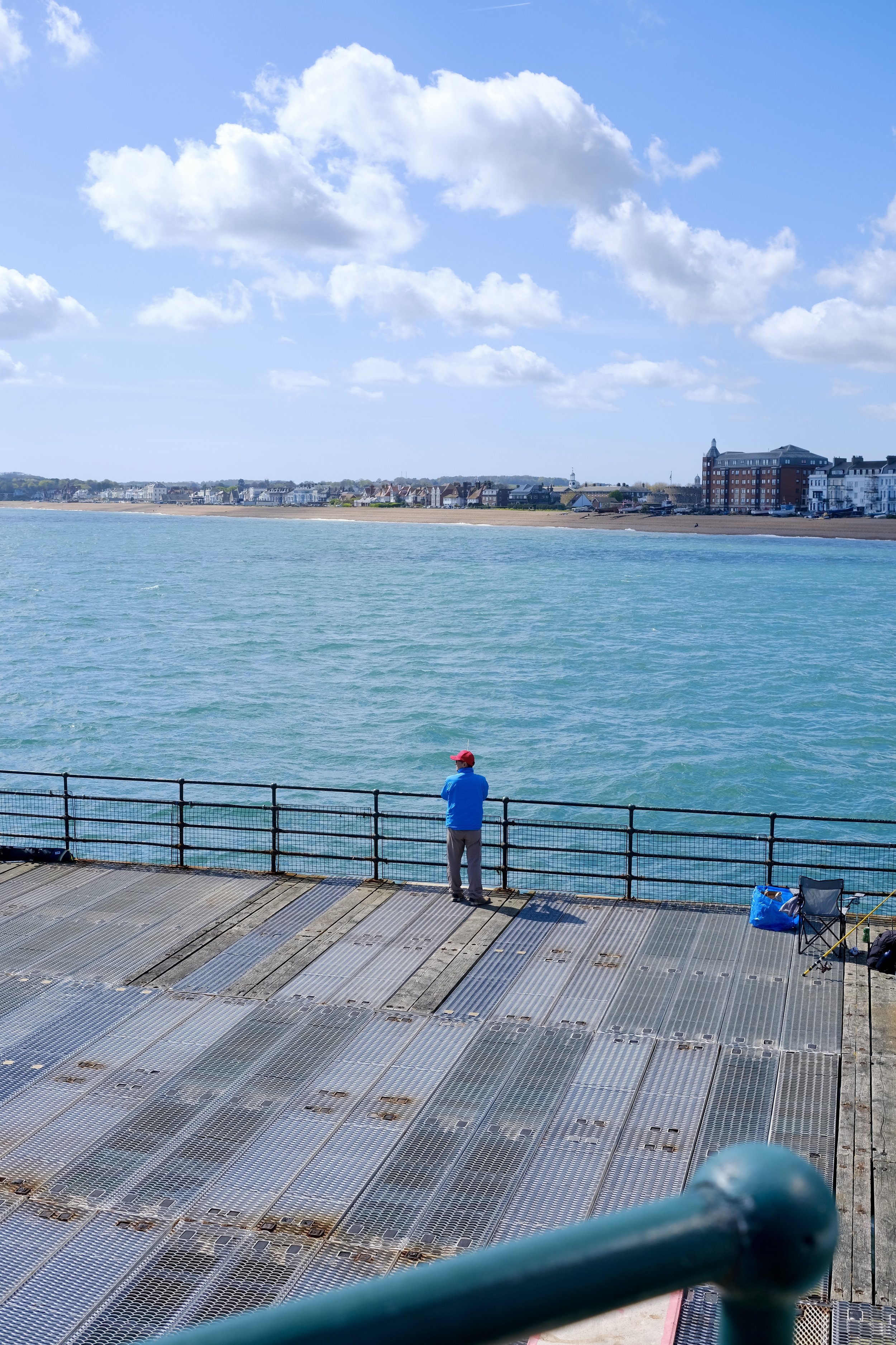 A person wearing a red cap and blue jacket standing on a boardwalk, looking out at the water with houses and buildings along the shoreline in the distance under a partly cloudy sky.