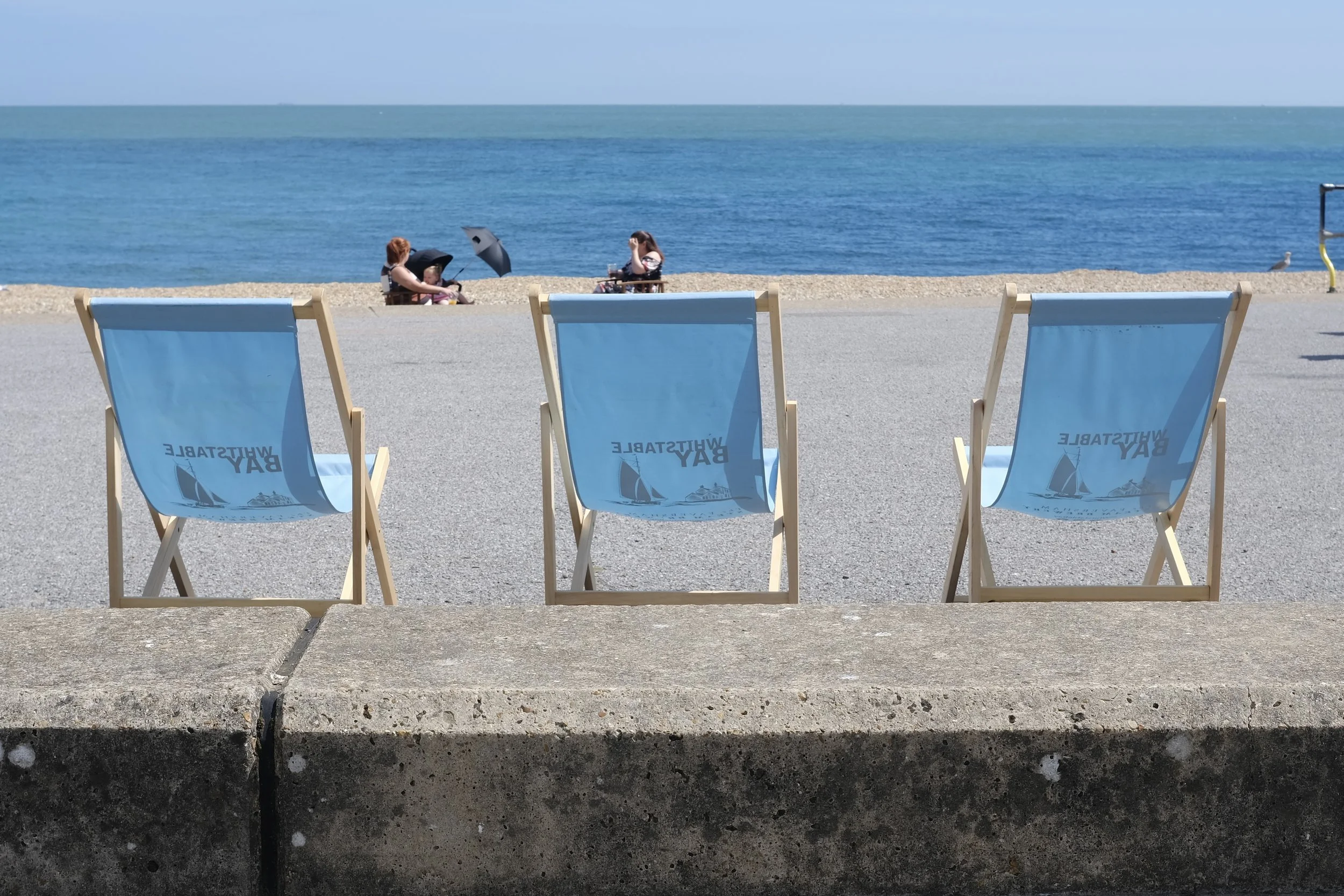 Beach scene with empty blue lounge chairs on a concrete surface, two people sitting near the shoreline with umbrellas, and a calm blue ocean in the background under clear sky.