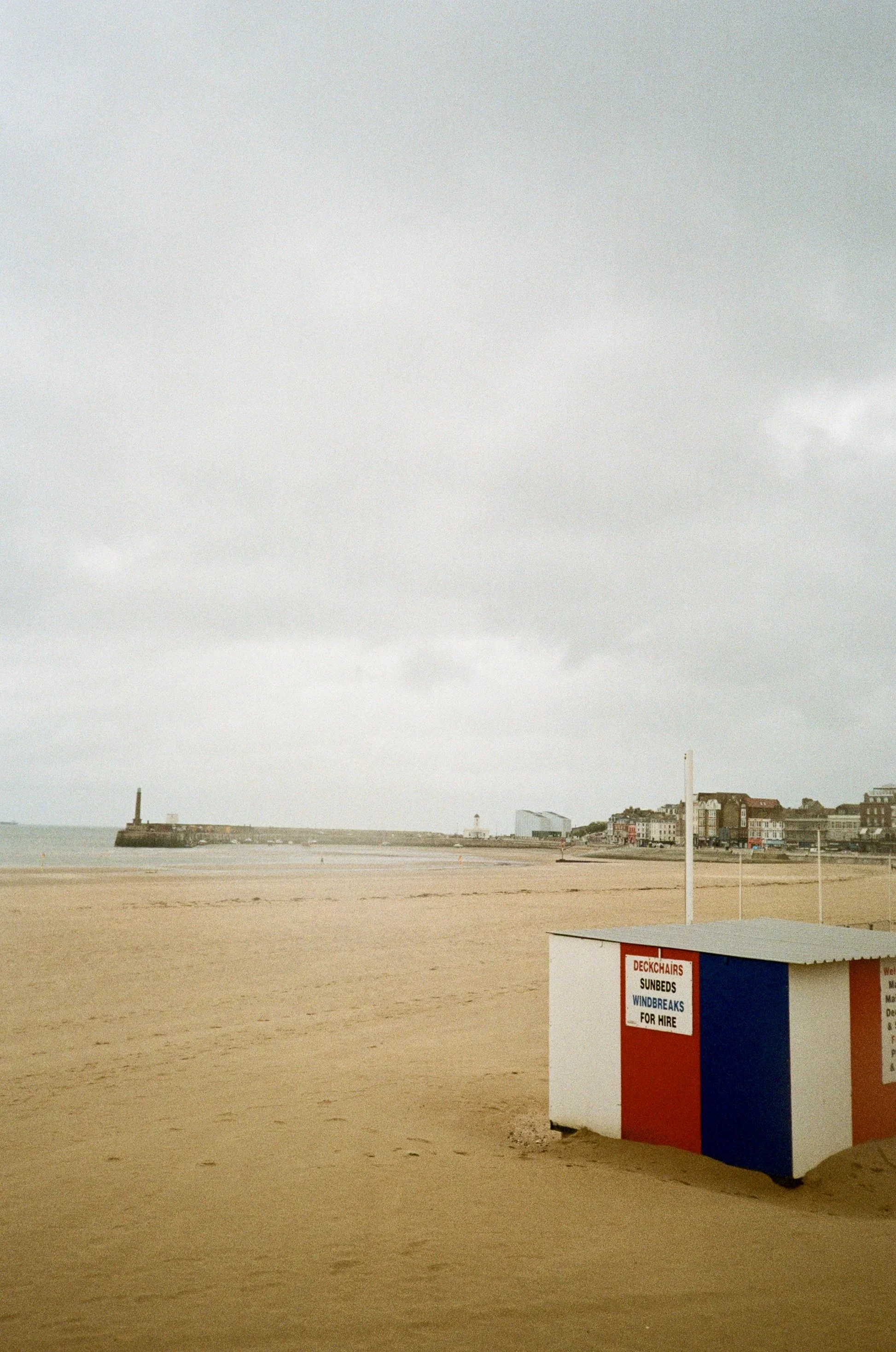A sandy beach with a small white and red building advertising deck chairs, sunbeds, windbreaks, and hire services. In the background, there is a harbor with a lighthouse on a jetty and a cityscape with buildings. The sky is overcast with gray clouds.