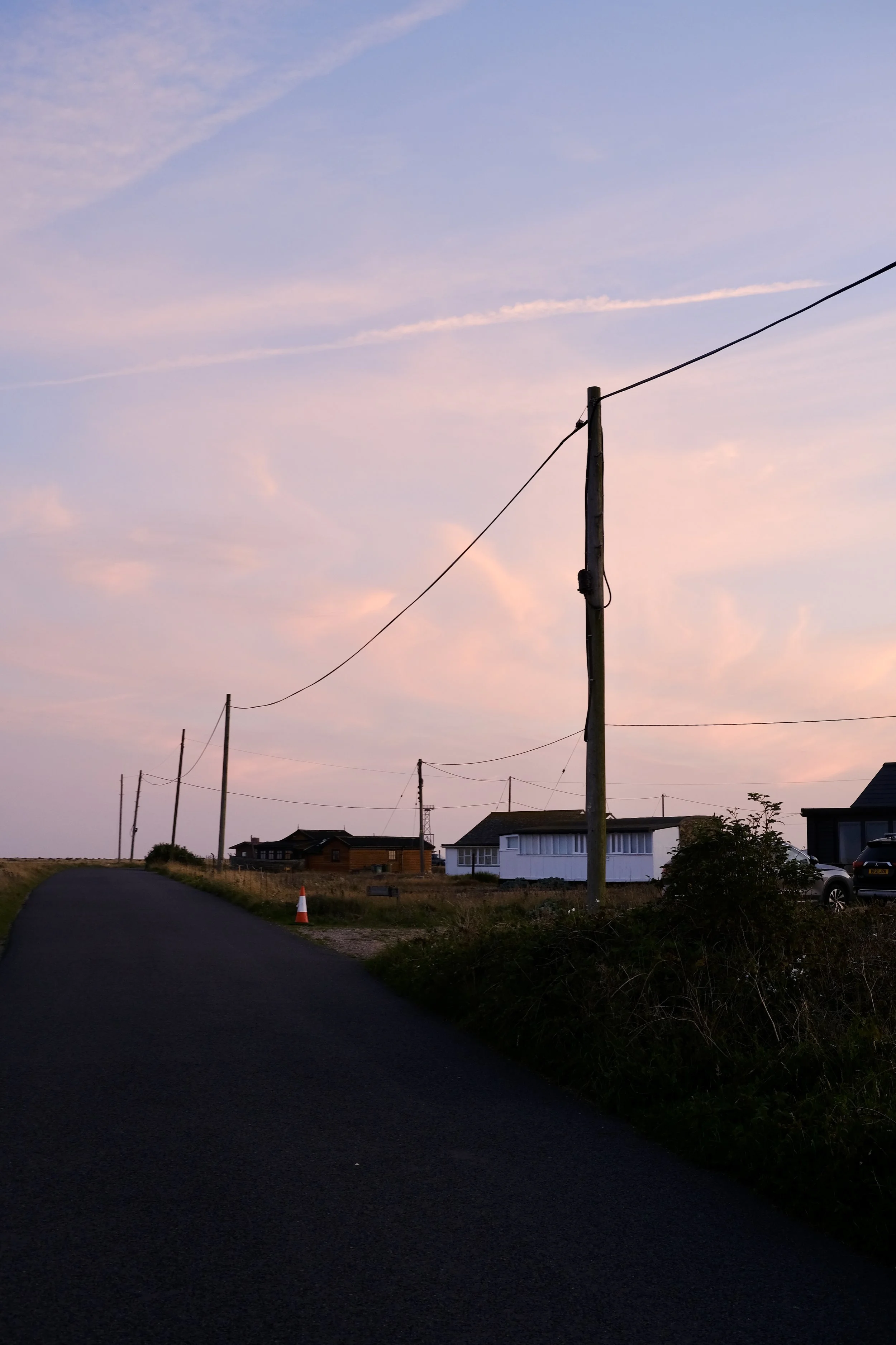 A rural street scene at sunset with a paved road, wooden utility poles with power lines, a few houses, a car, and a traffic cone, under a pastel sky with clouds.